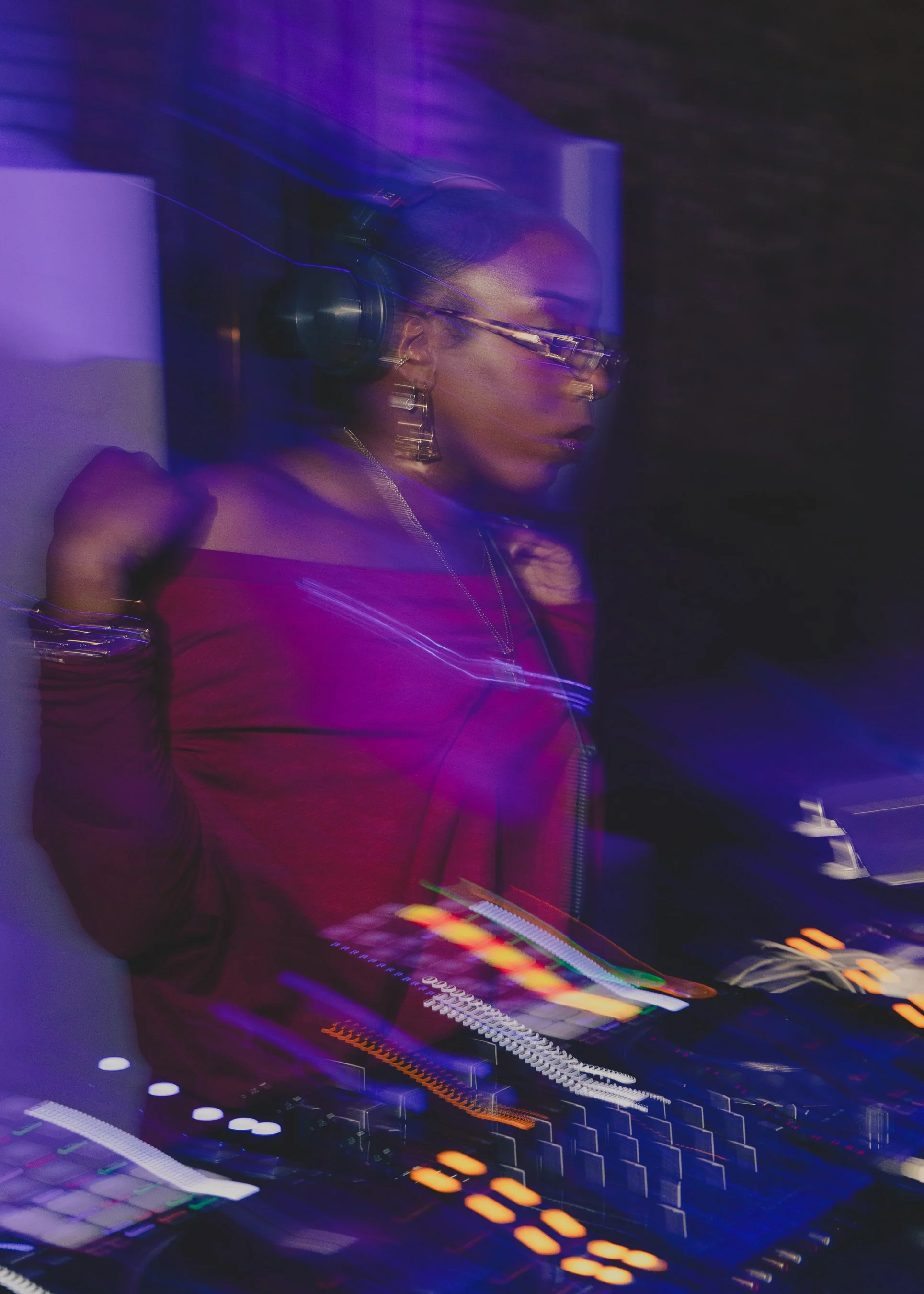 A female DJ wearing headphones and glasses, performing with a DJ mixer and turntables in a dark, colorful, and vibrant environment with purple and orange lights.