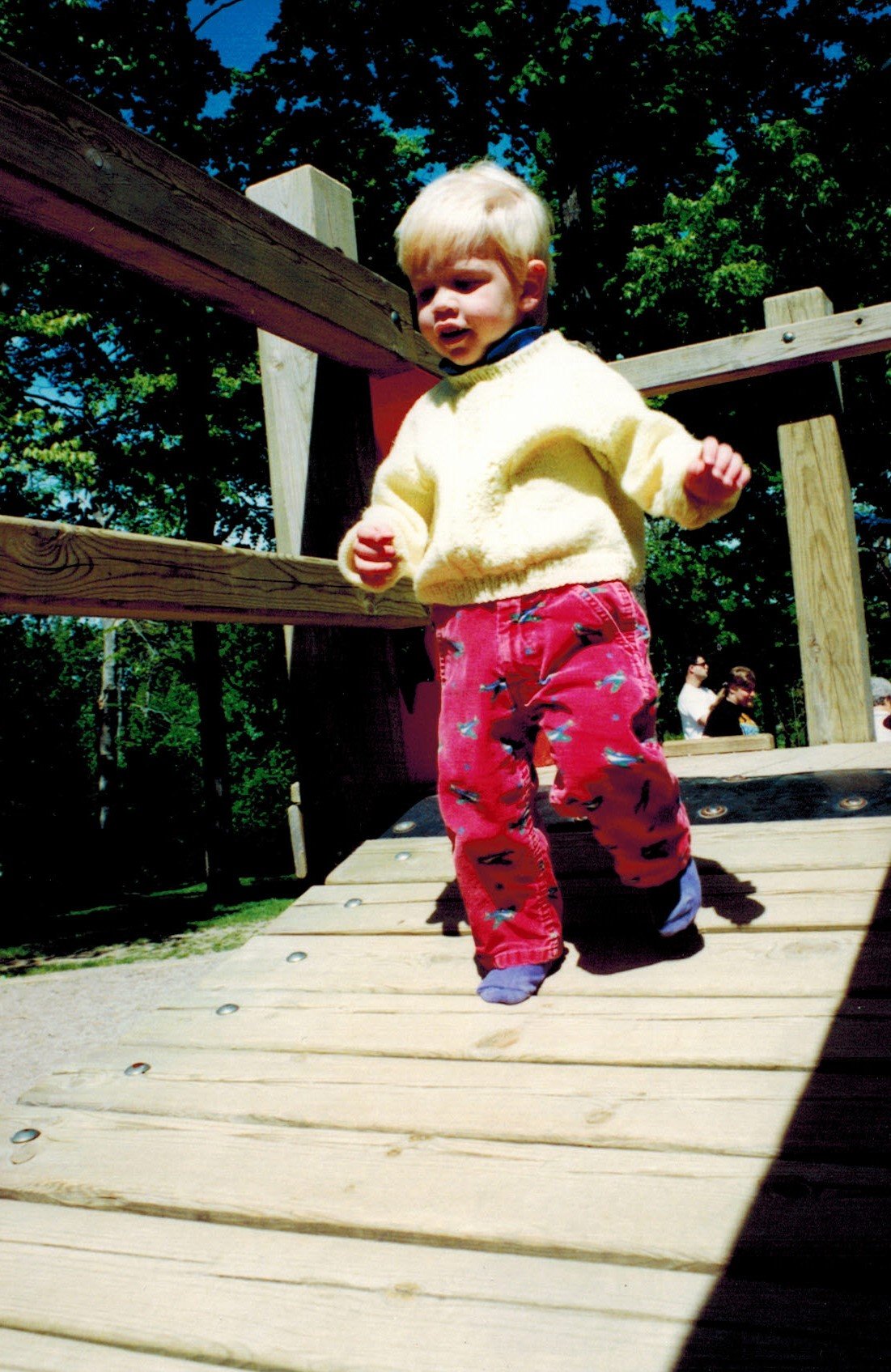 Young child with blonde hair wearing a yellow sweater and red pants walking on a wooden bridge at a park on a sunny day, with green trees and a few adults in the background.