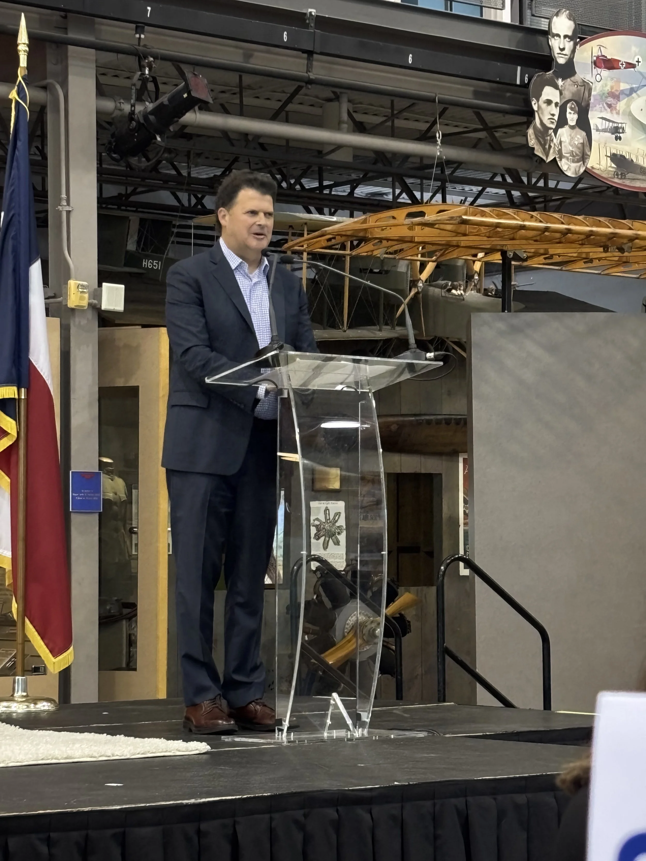 A man in a suit speaking at a podium during a presentation or speech at a museum or exhibition space with historical aircraft and portraits in the background.