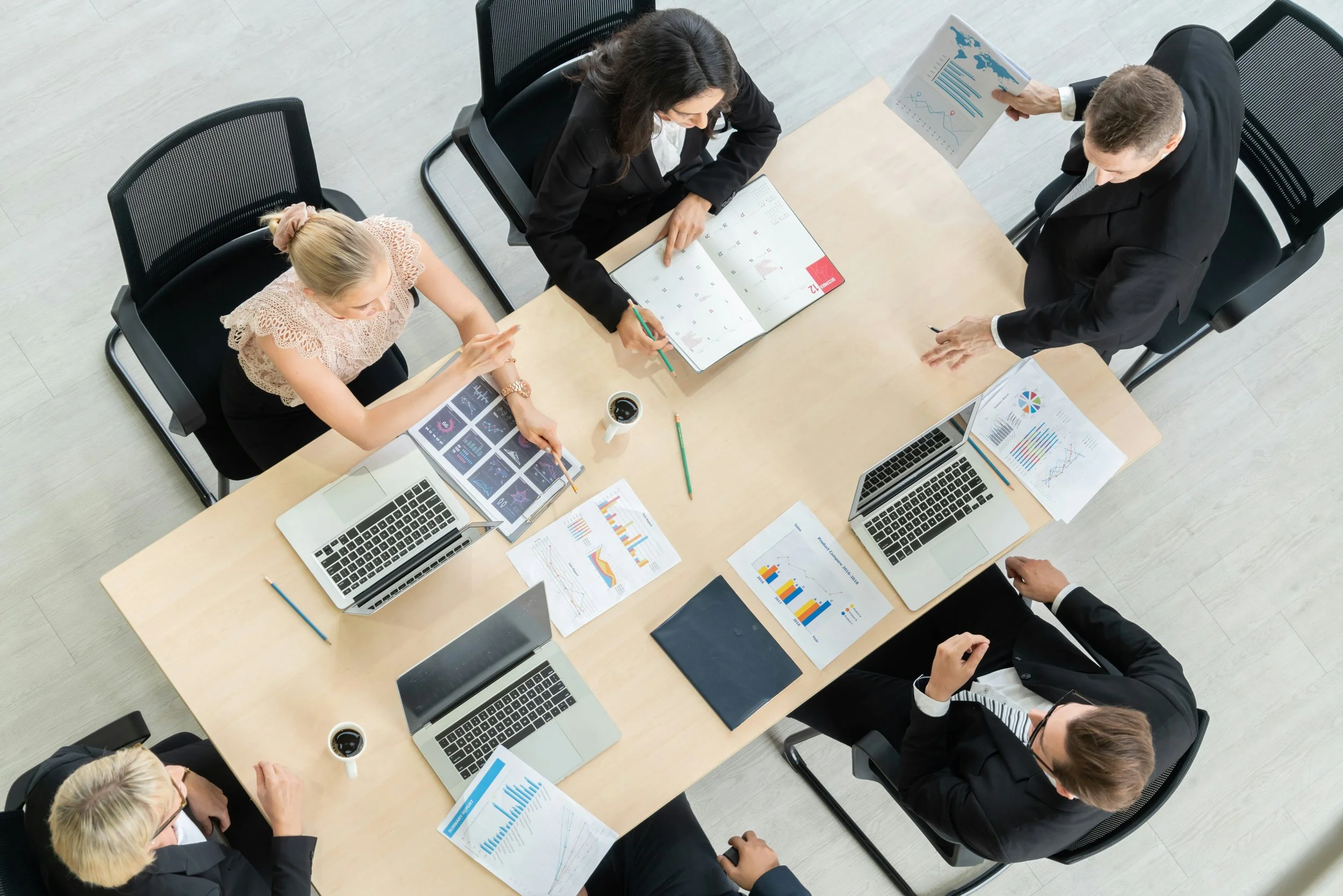 Top-down view of five business professionals in formal attire sitting and standing around a wooden conference table with laptops, printed charts, graphs, and coffee cups, engaged in a meeting.