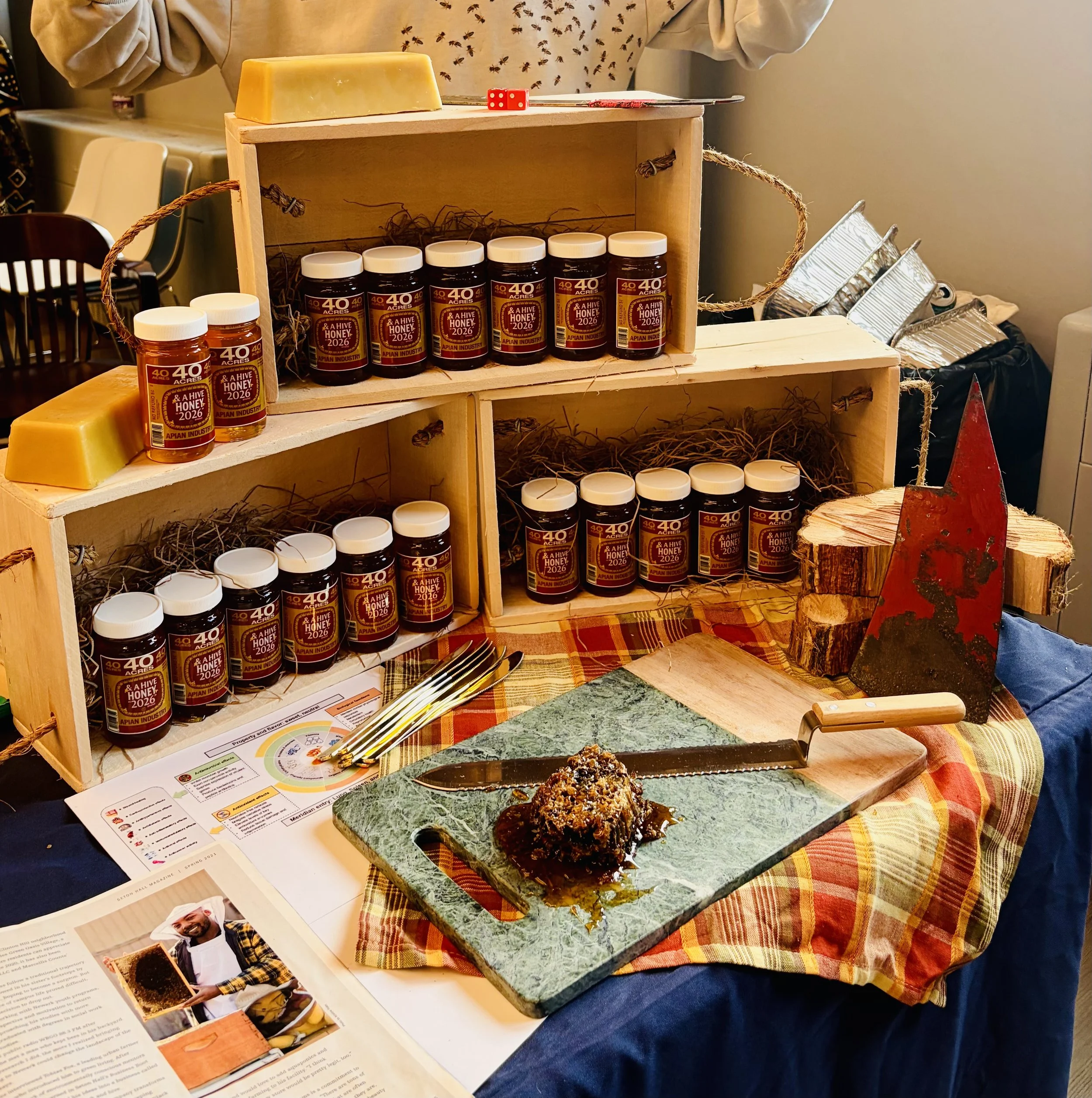 Jars of honey labeled as '40 Acres & A Hive Honey 2026' arranged on a wooden shelf. A block of yellowish soap, a red die, and some aluminum containers are also on the shelf. In the foreground, a knife with a green and gray handle and a piece of honeycomb on a green tray are visible, placed on a table with a red plaid tablecloth.