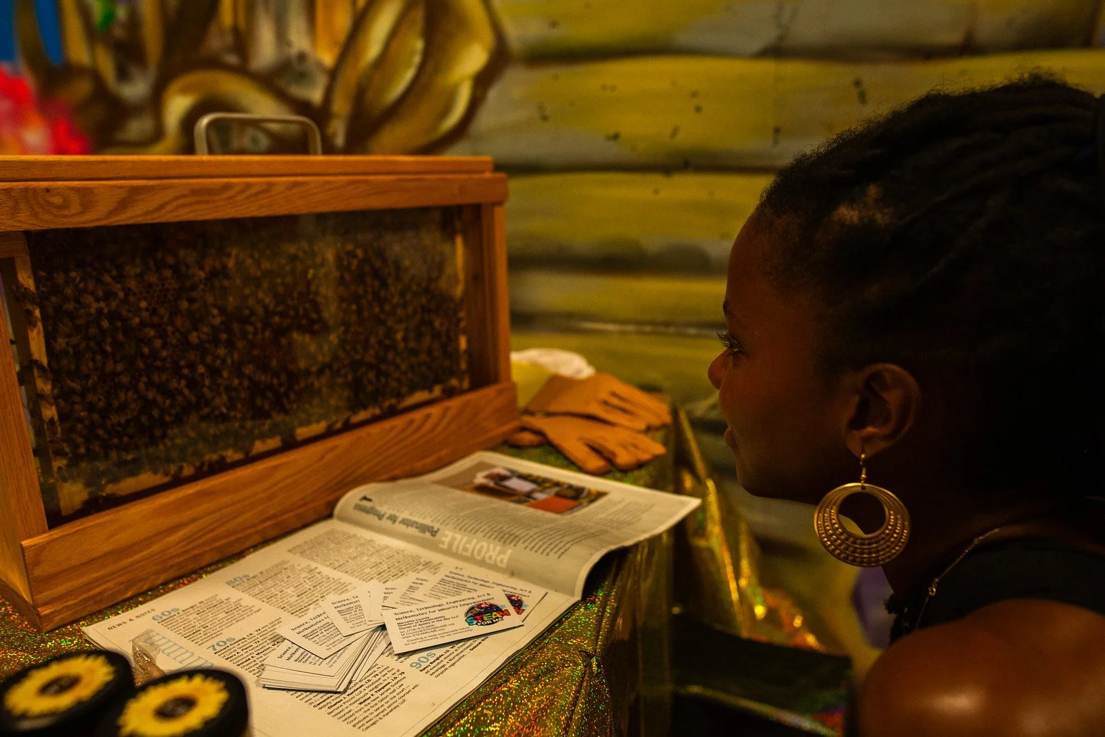 A woman with braided hair and large hoop earrings looks at a display of beekeeping frames and an open magazine on a glittery table, with a wooden beehive box containing bees nearby.