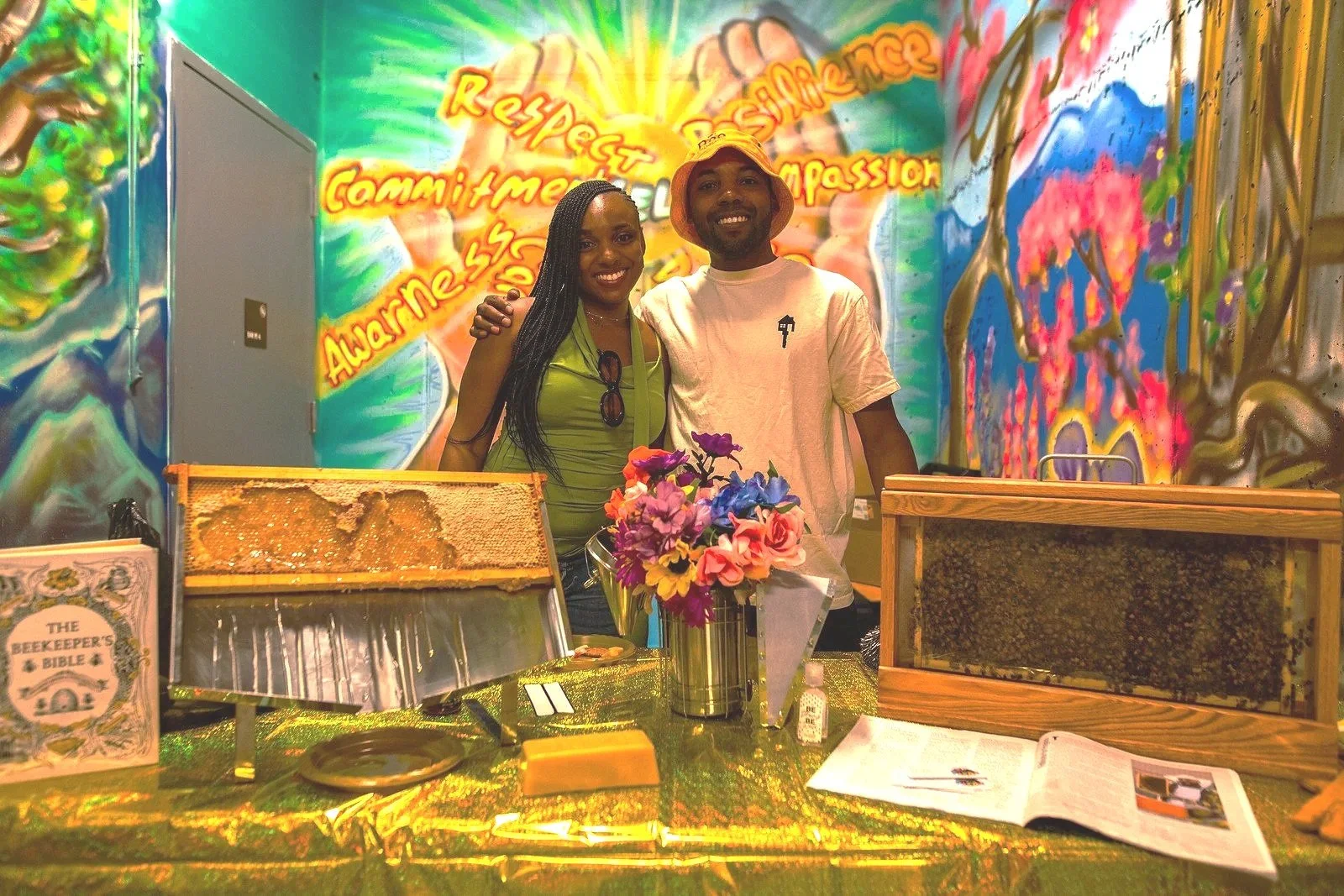Family founders standing behind a table with honey and beekeeping supplies, in front of a colorful mural with words like 'Respect,' 'Compassion,' and 'Awareness'.