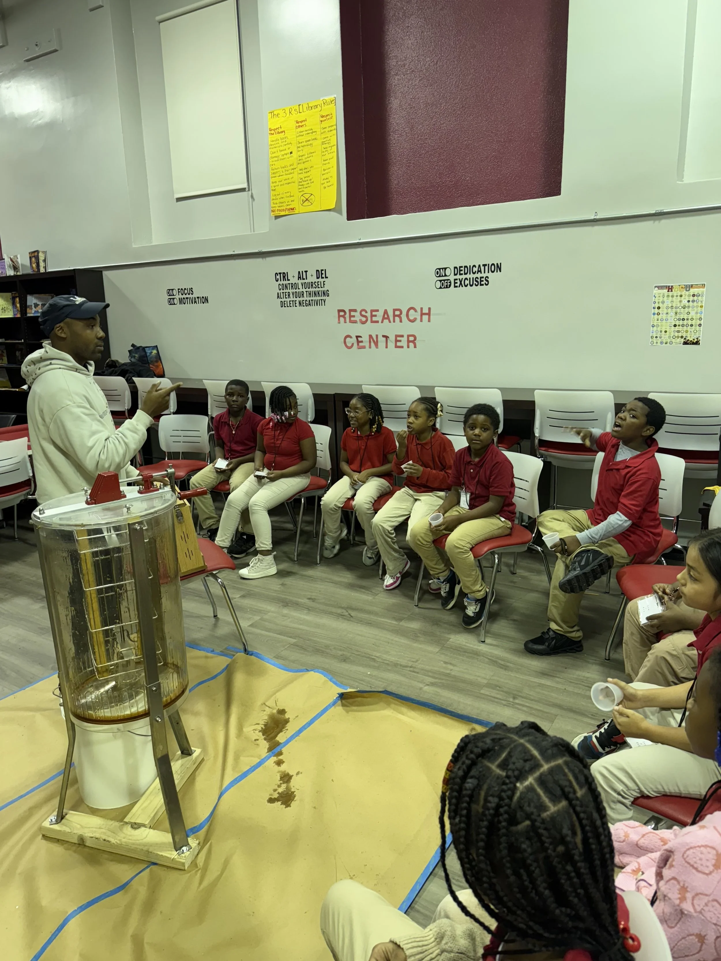 Children seated in chairs during a science demonstration at a research center, with a scientist explaining and a large science apparatus nearby.