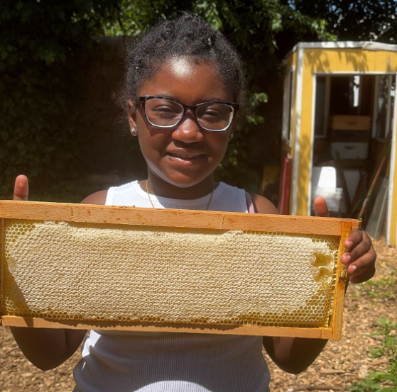 Young girl with glasses holding a honeycomb frame outdoors.