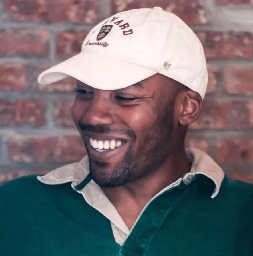 Founder Marcellis Counts wearing a white baseball cap and a dark green shirt with a beige collar, against a brick wall background.