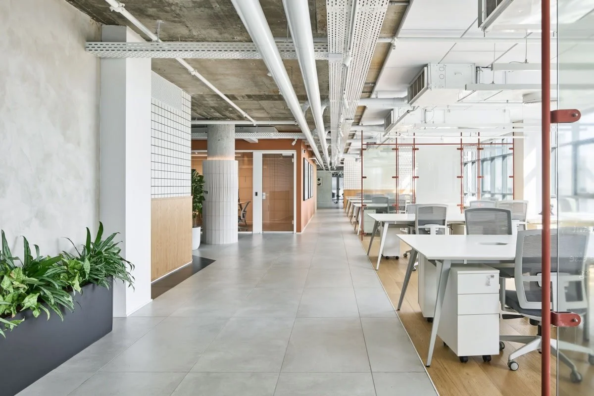 Open-space d'ufficio moderno con scrivanie, sedie, pareti di vetro e piante decorative, con un design industriale e illuminazione naturale.