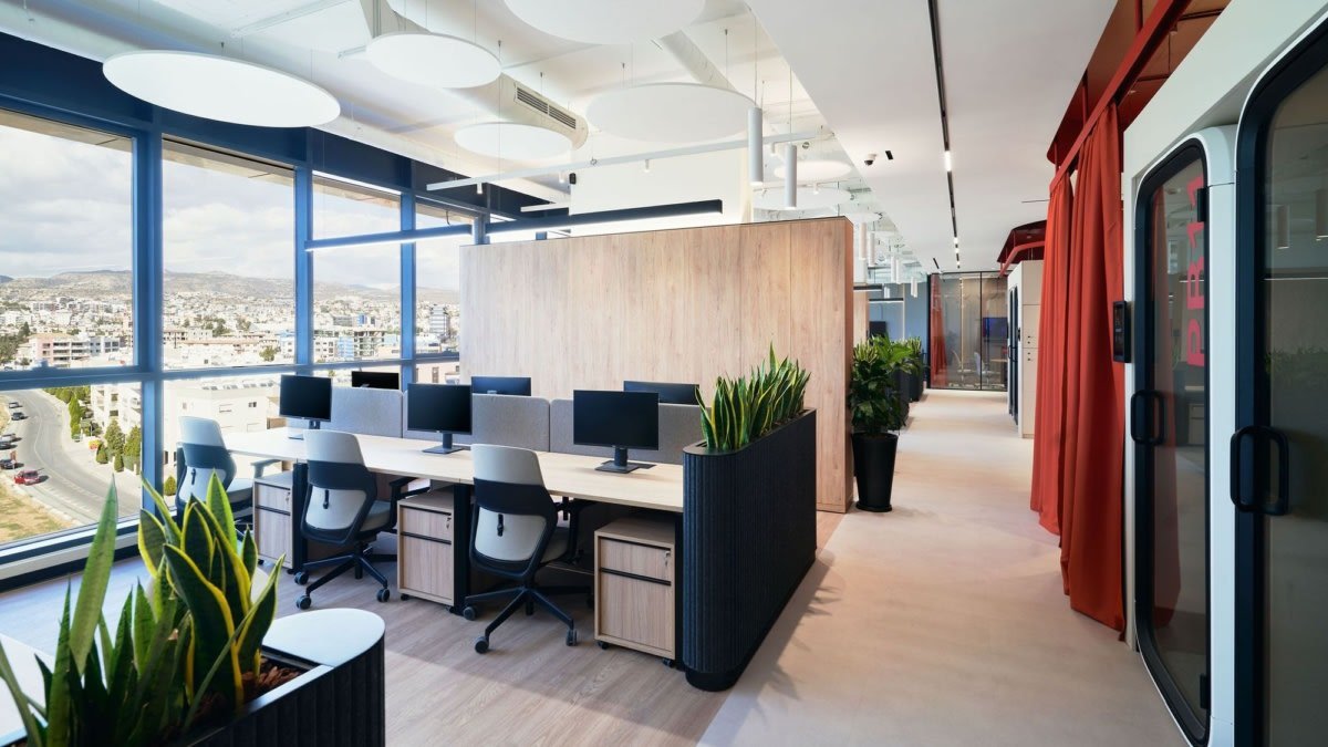 Spazio di coworking con postazioni di lavoro, piante verdi e grandi finestre che offrono vista sulla città.
