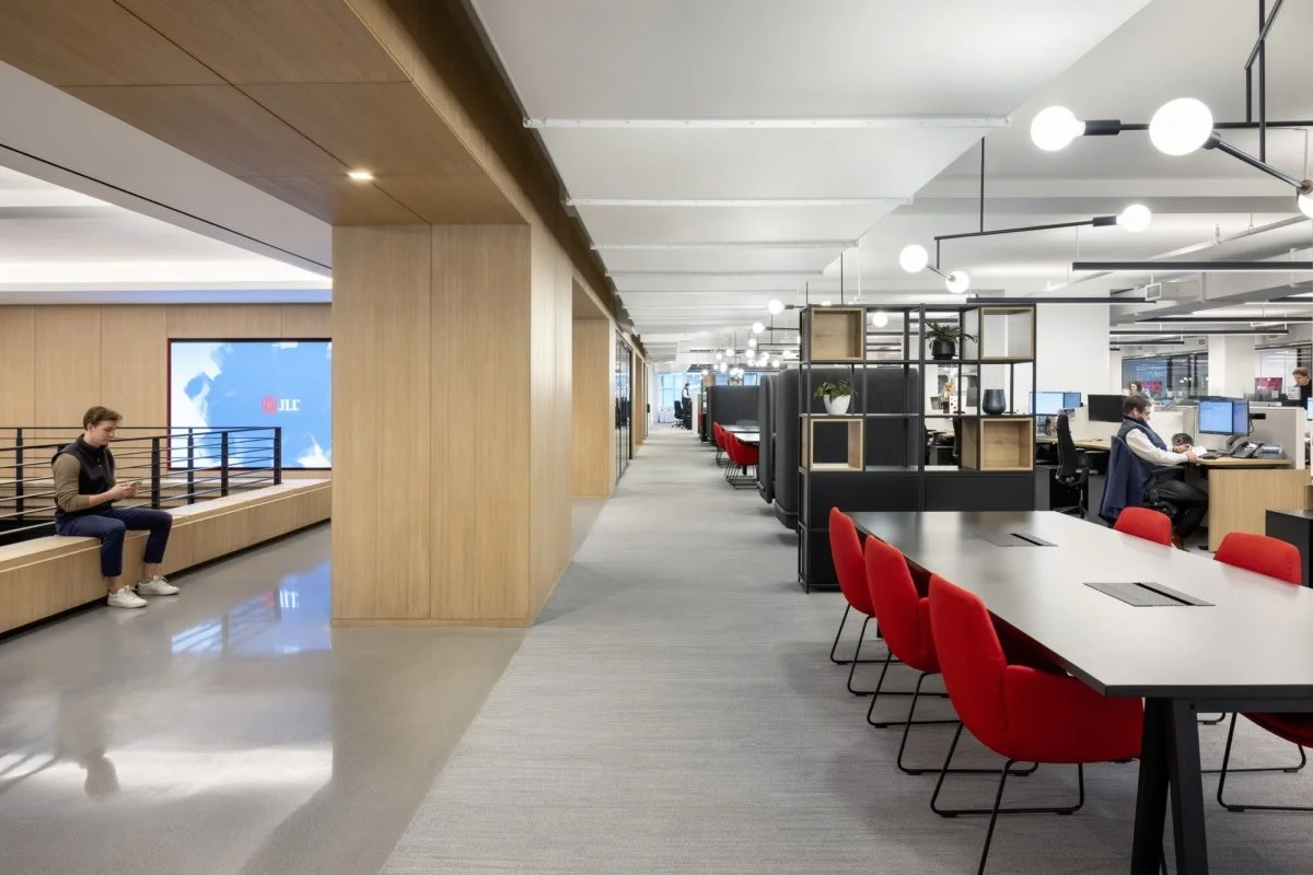Interno di un ufficio moderno con postazioni di lavoro, sedie rosse, pareti di legno e illuminazione a soffitto. Una persona lavora al computer e un'altra aspetta seduta.