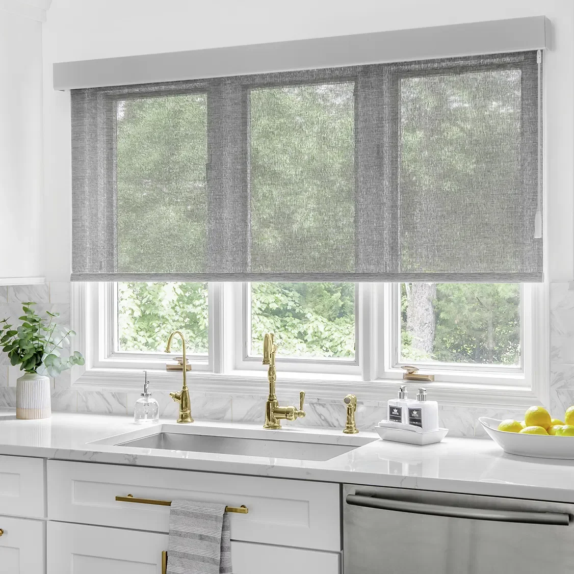 Modern kitchen with white marble countertops, gold fixtures, a gray roller shade window covering, a potted plant, and a bowl of lemons.