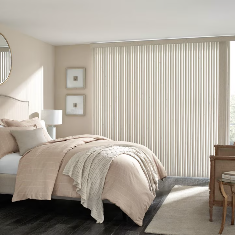 A bedroom with a neatly made bed with beige and cream bedding, a nightstand with a white lamp, framed artwork on the wall, a large window with vertical blinds, and a wooden chair with books on a small table.