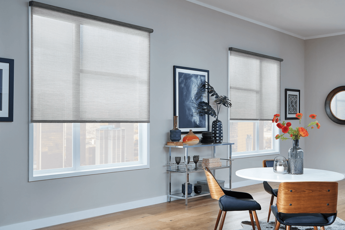 Living room with two large windows covered by beige roller blinds, a small table with a vase and flowers, a bookshelf with decorative items, and a round table with chairs in a modern apartment.