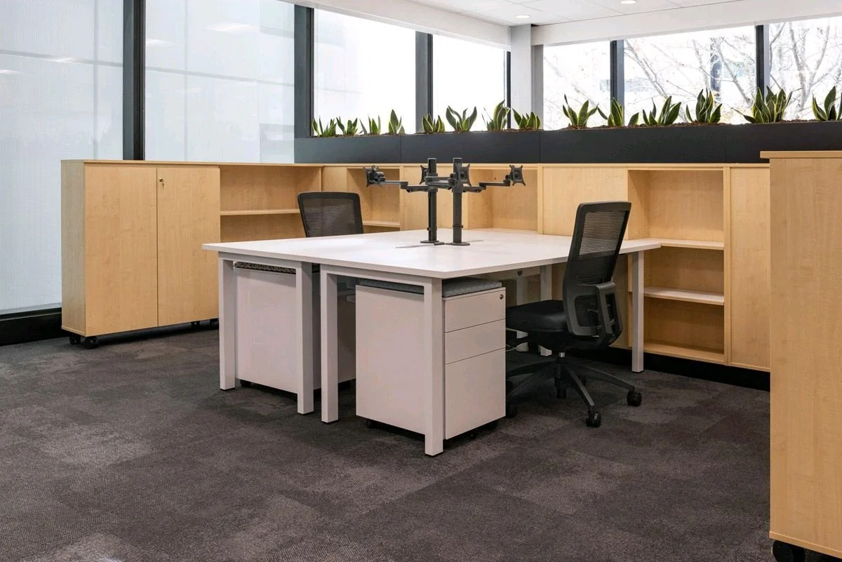 Empty office workspace with a white desk, black office chair, and a drone on the desk, surrounded by wooden cabinets and a row of potted plants near large windows.