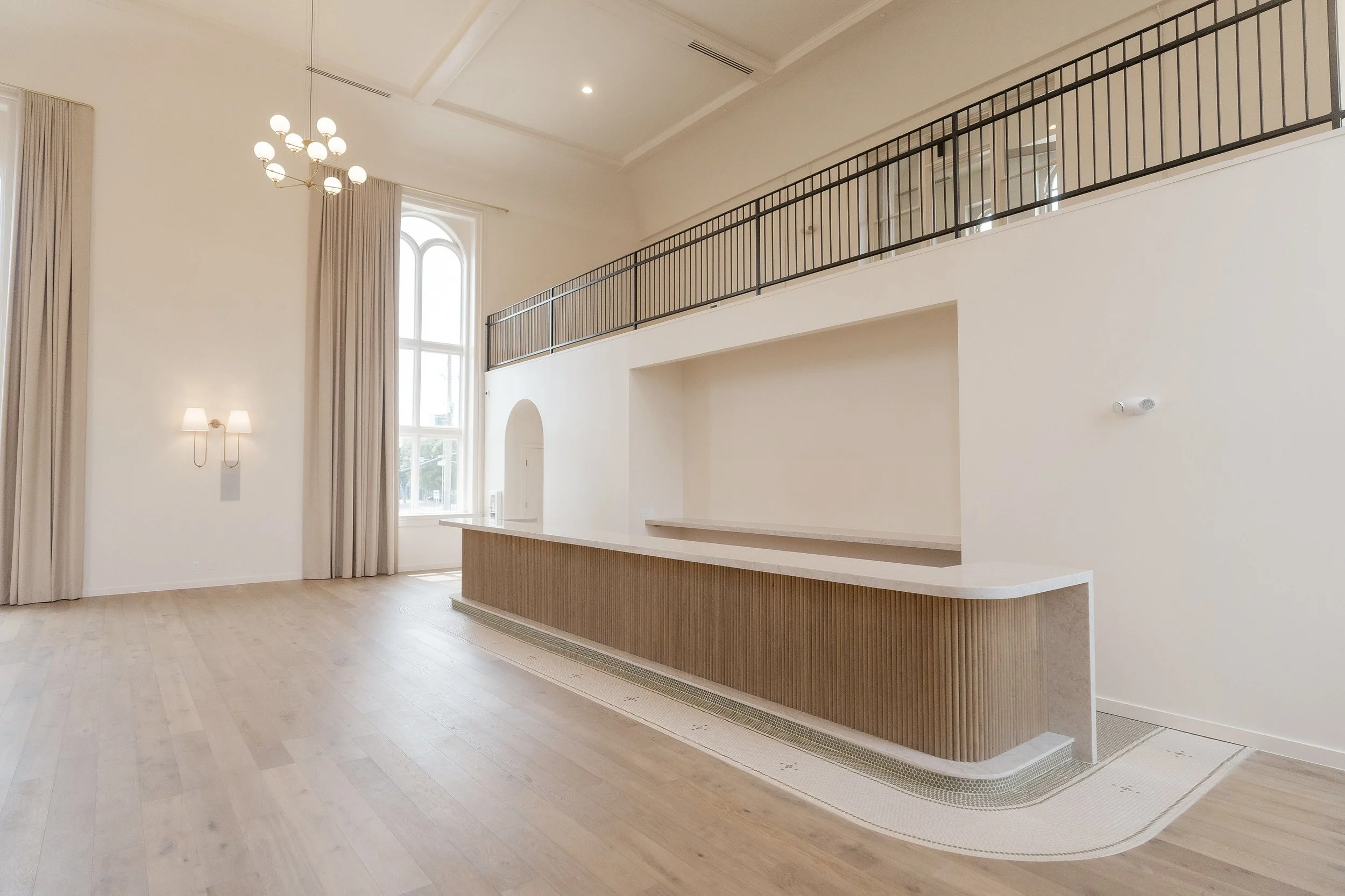 Interior of a spacious, modern room with high ceilings, large arched windows, a chandelier, wall-mounted lamps, and a balcony with a metal railing.