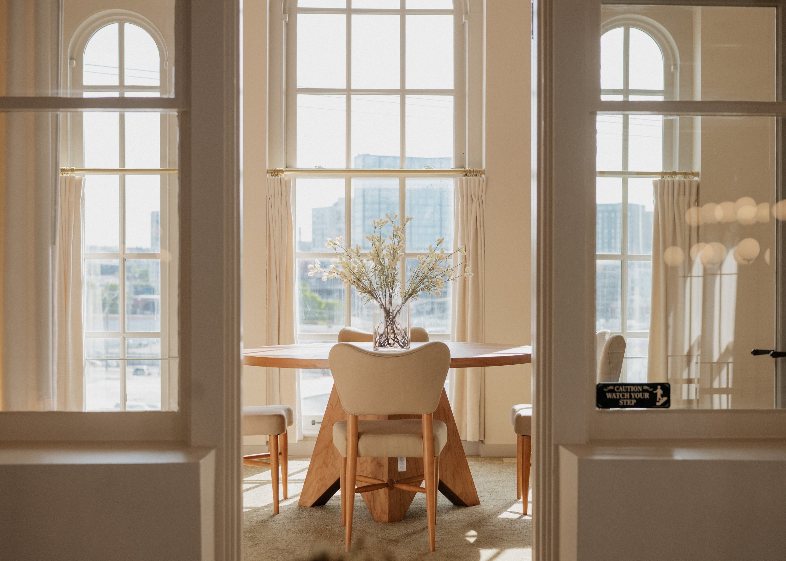View through a set of glass-paneled doors into a bright room with a round wooden dining table, white chairs, a glass vase with flowers, large windows with curtains, and a cityscape in the background.