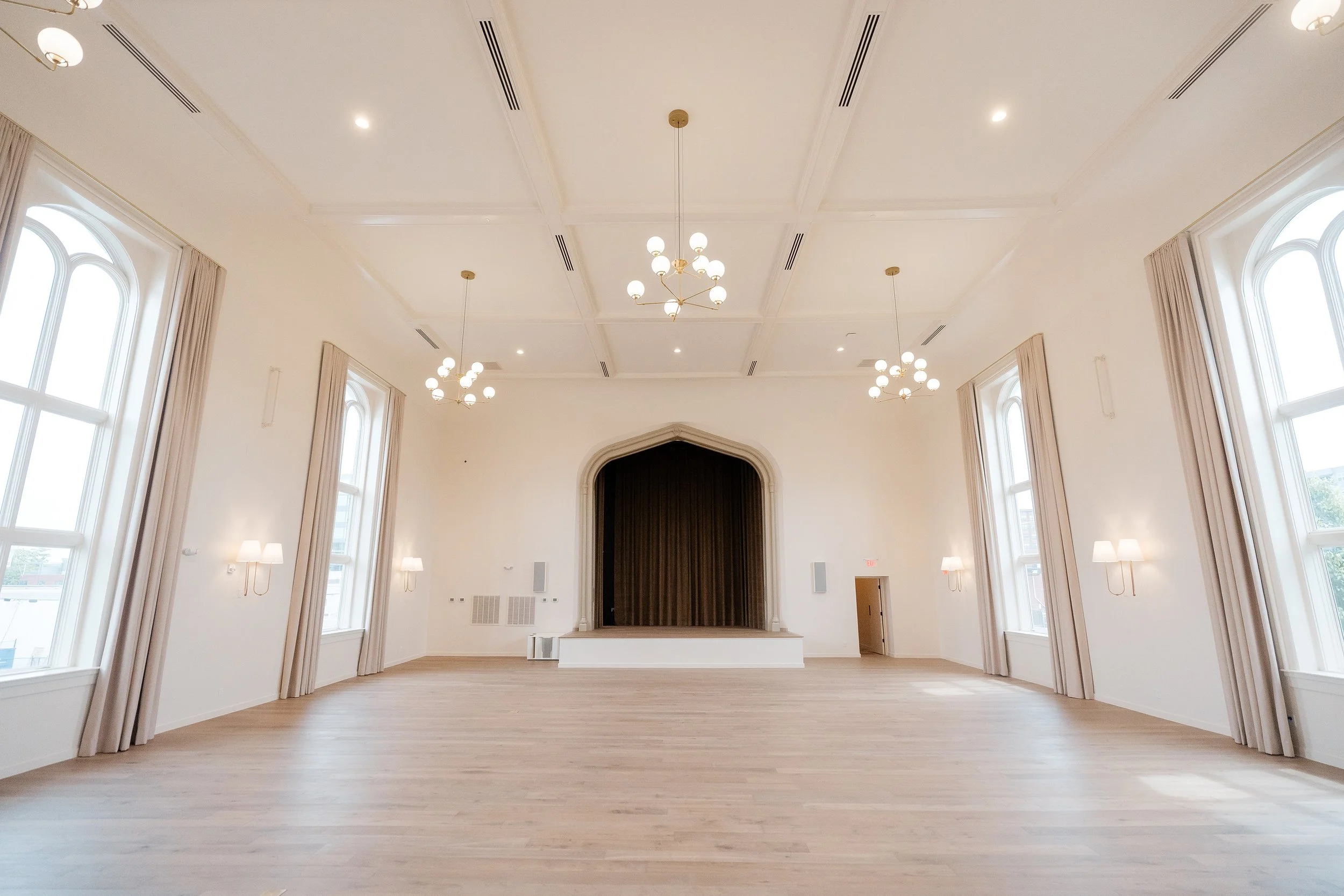 A spacious, empty room with high ceilings, light-colored walls, large arched windows with curtains, and modern chandeliers.