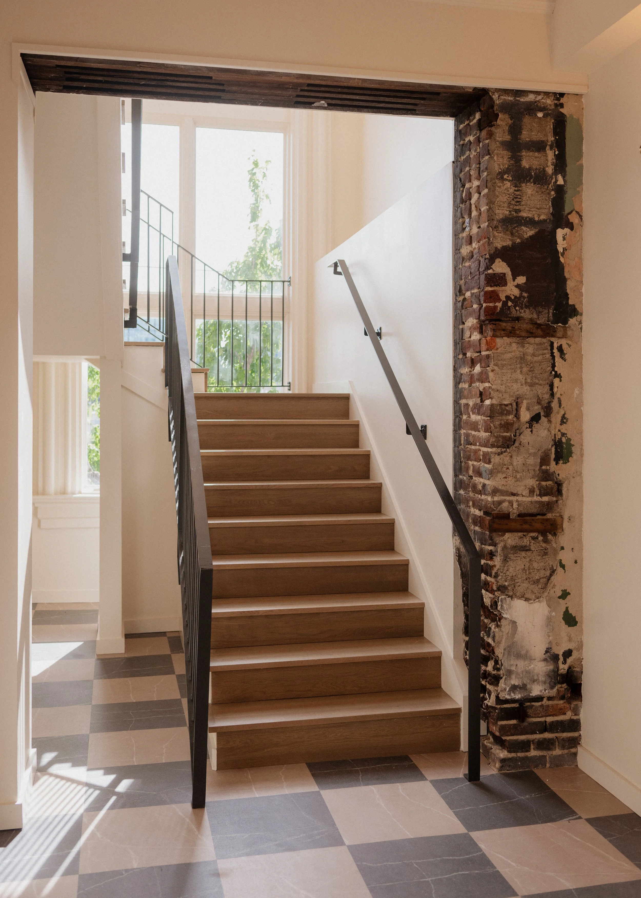 Indoor staircase with wooden steps, black metal railing, exposed brick wall on the right, large window at top, sunlight casting shadows on checkered tile floor.