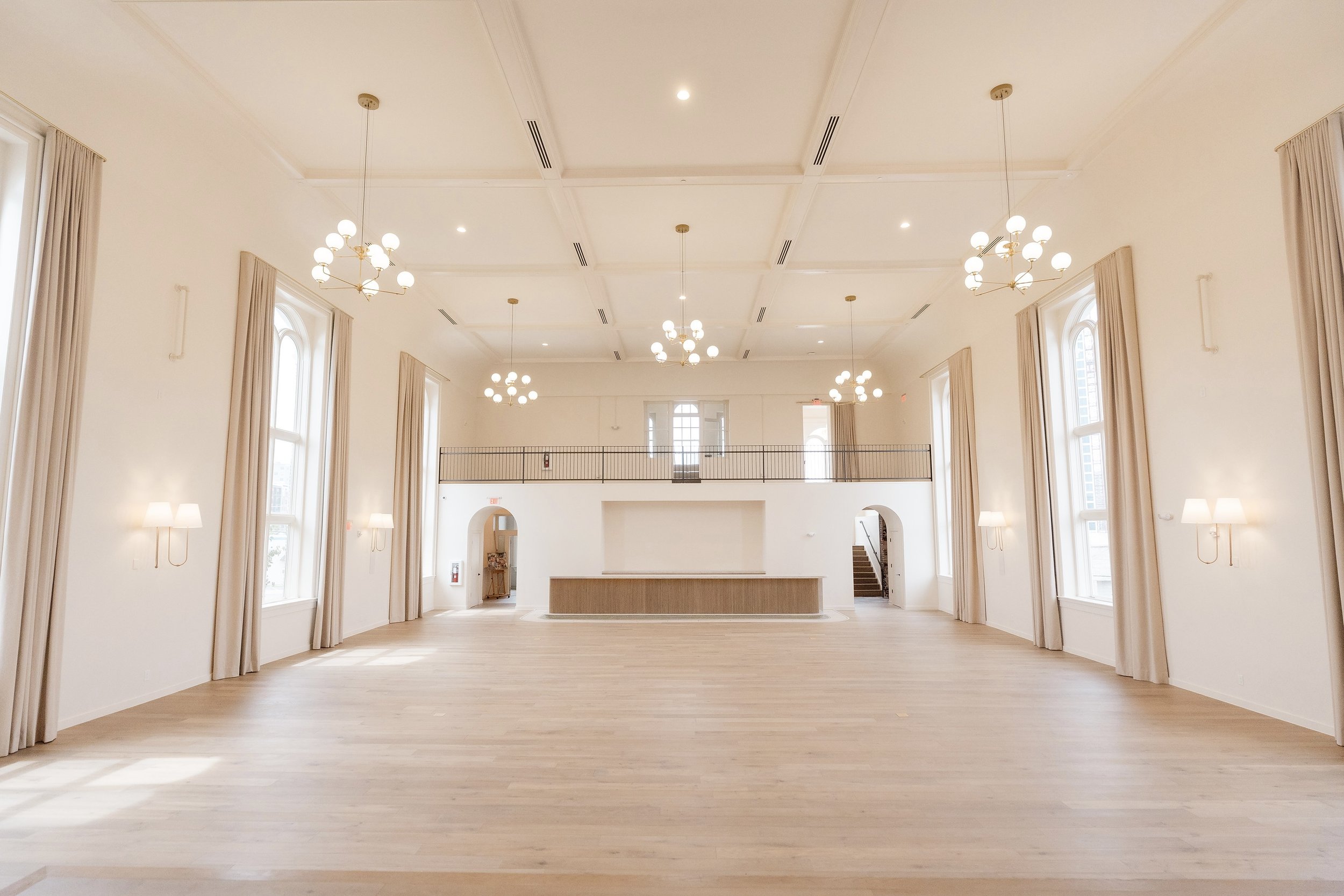 A spacious, well-lit room with high ceilings, large windows with curtains, and multiple chandeliers with globe lights. The room has light-colored wooden flooring and a small balcony with a railing at the far end.