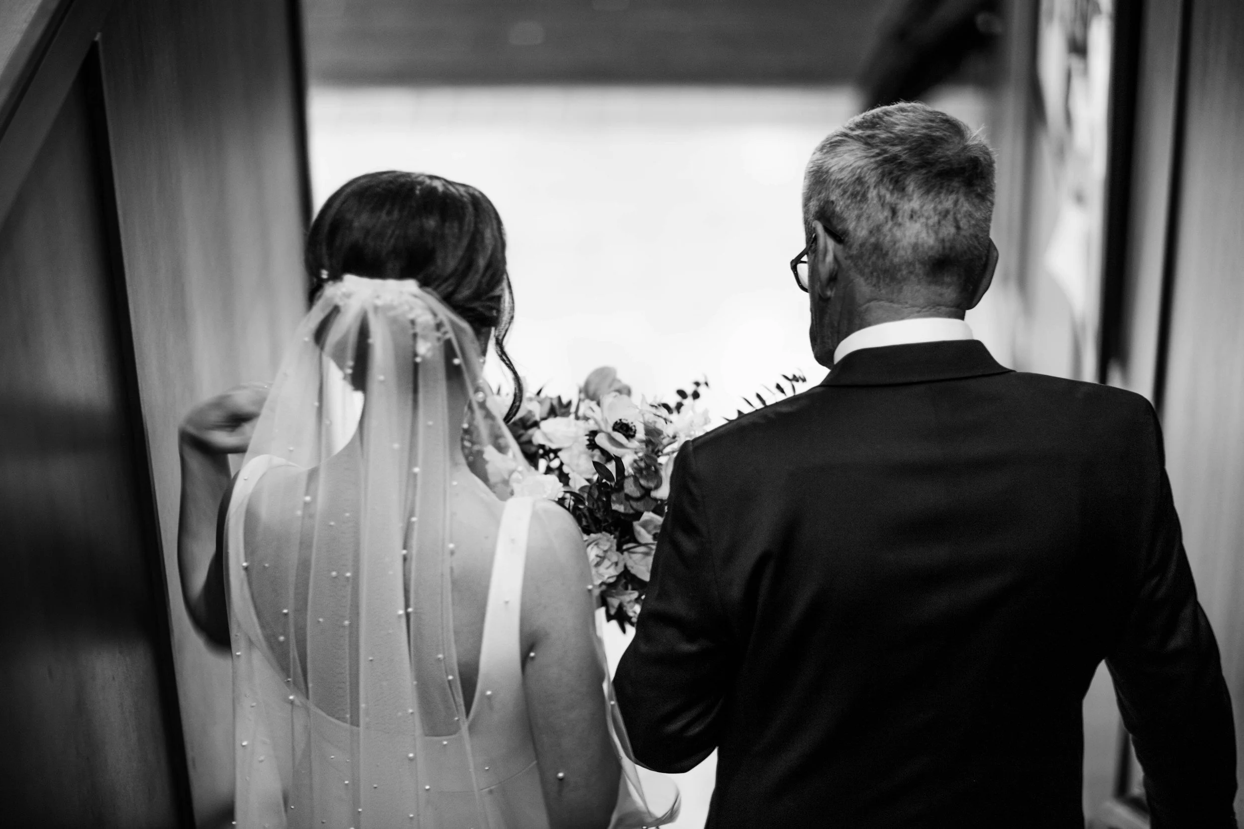 A groom in a black tuxedo holding hands with a bride in a strapless wedding dress and tartan sash, standing against a brick wall.