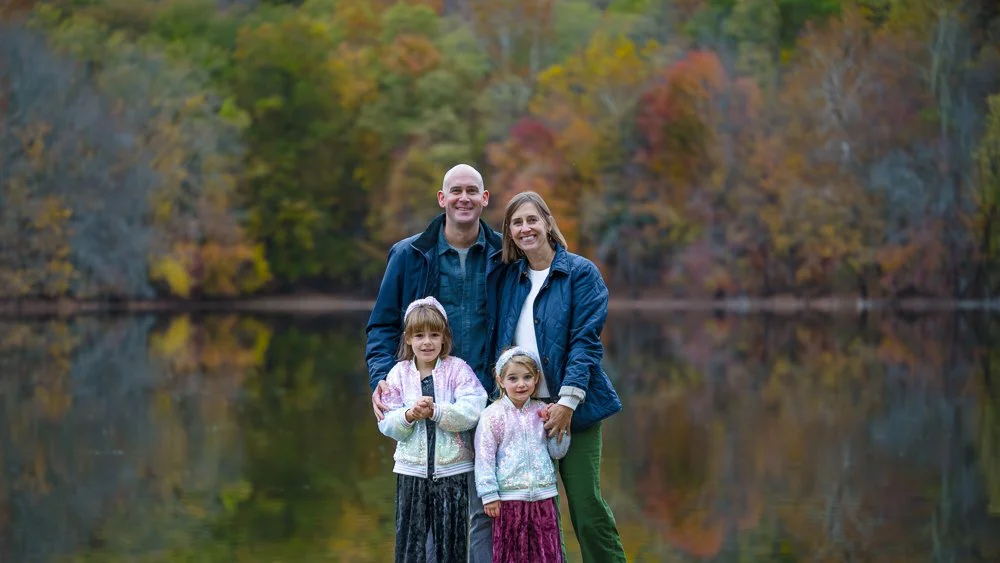 A family of four standing by a lake with colorful autumn trees in the background.