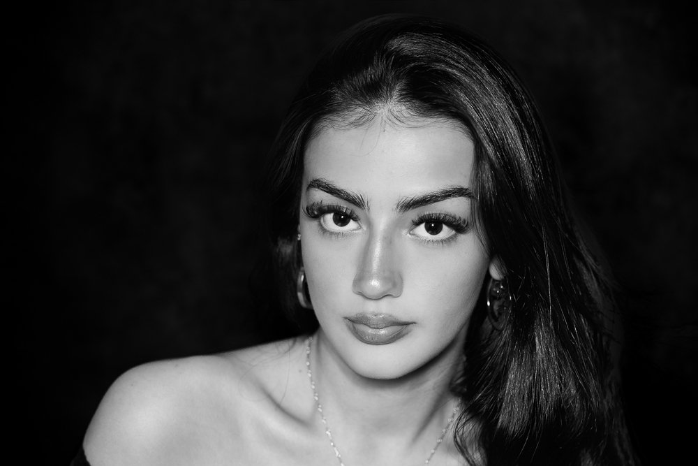 Close-up profile of a young woman with dark hair and fair skin, outdoors with blurred greenery in the background.