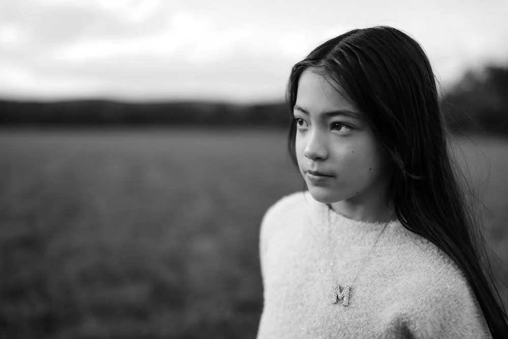 Black and white photo of a young girl with long hair standing outdoors in an open field, looking to the side.