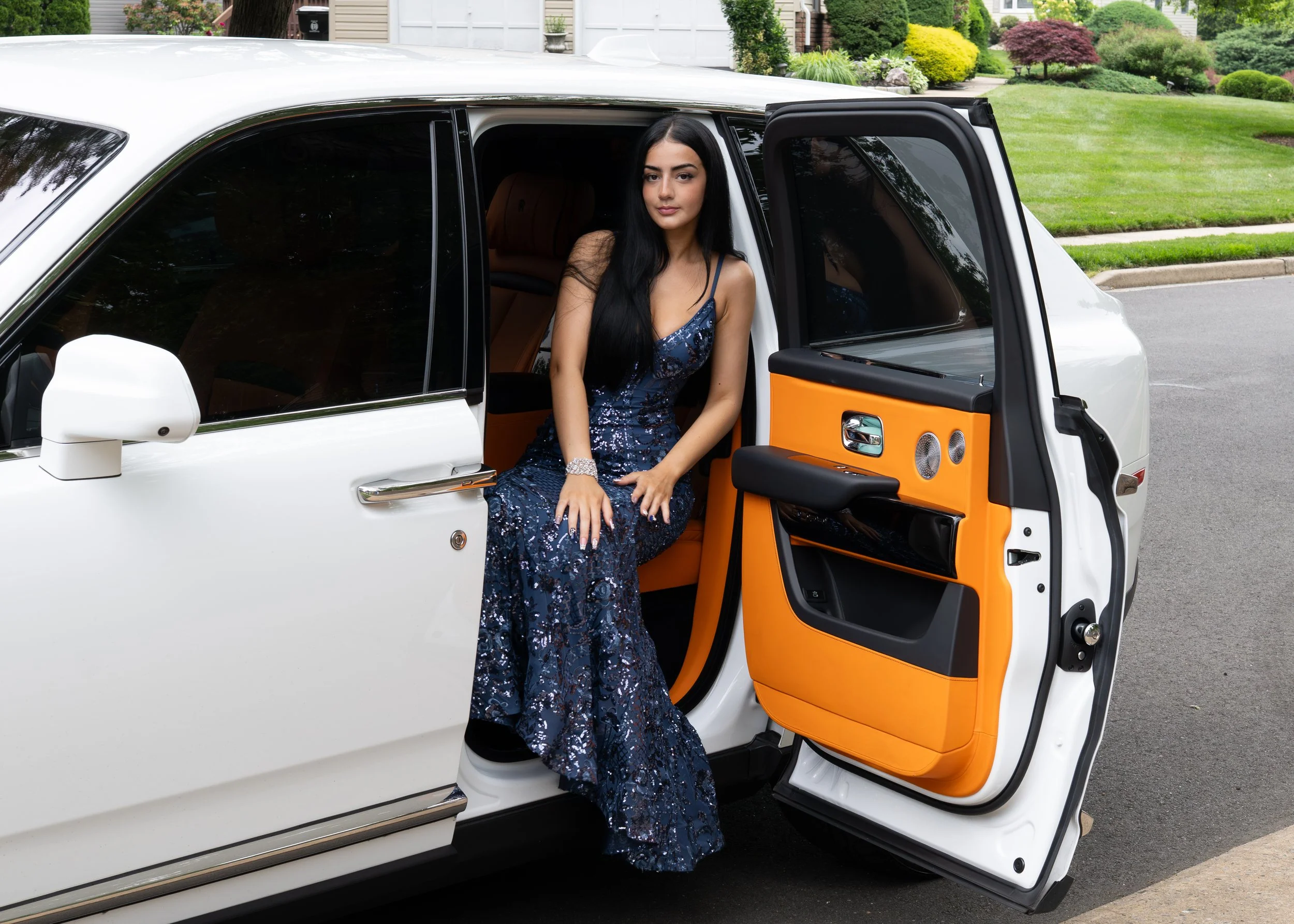 A young woman in a dark blue, sequined gown sitting in the open door of a white luxury SUV with orange and black interior, on a suburban street with well-maintained lawns and gardens in the background.