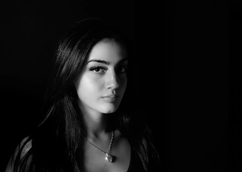 Side profile of a young woman with dark hair and natural makeup against a blurred green background.