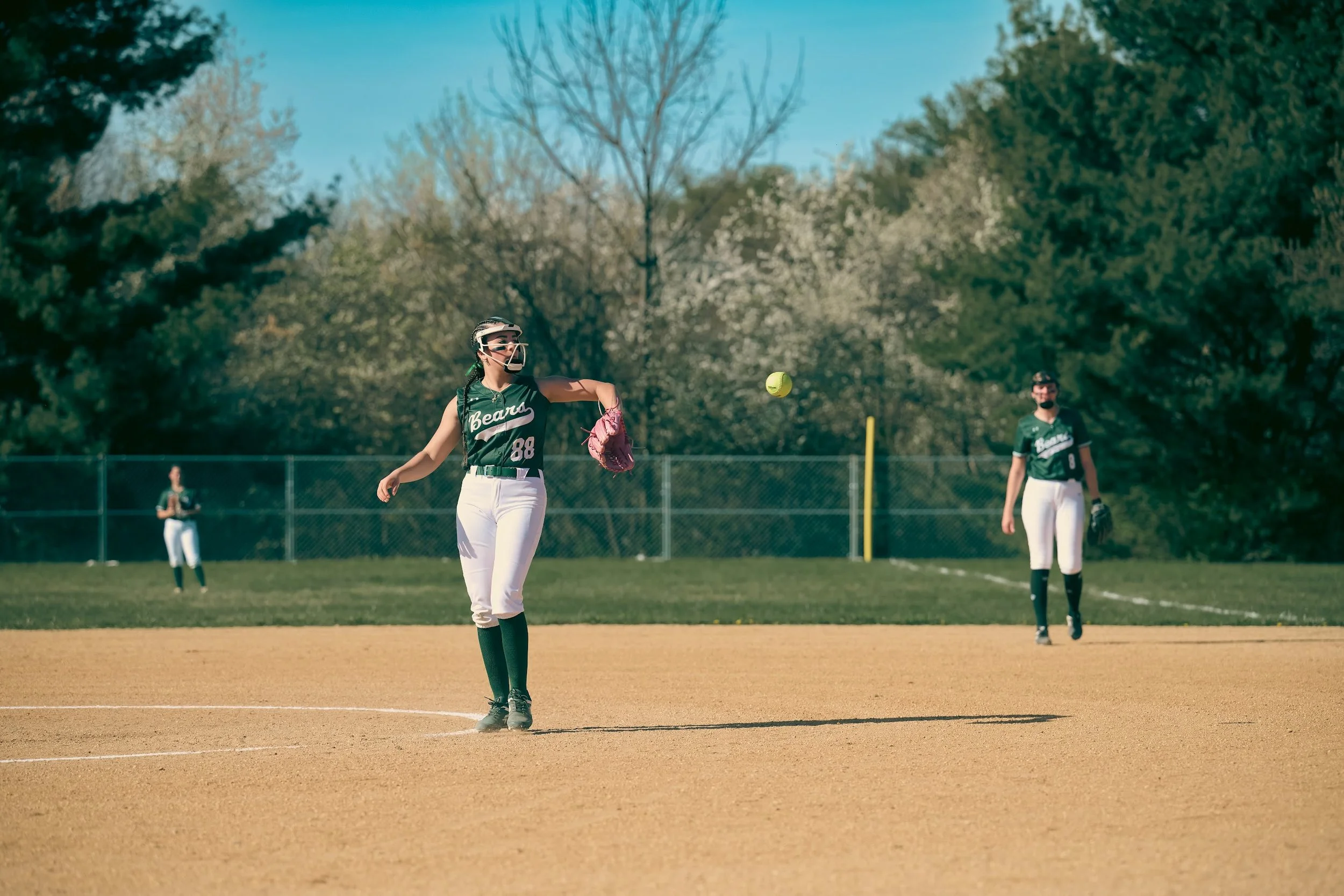 A female softball player in a green uniform with the number 88, wearing a face guard and glove, is actively throwing a softball on the field. Two other players in similar uniforms are visible in the background, with trees and a chain-link fence surro