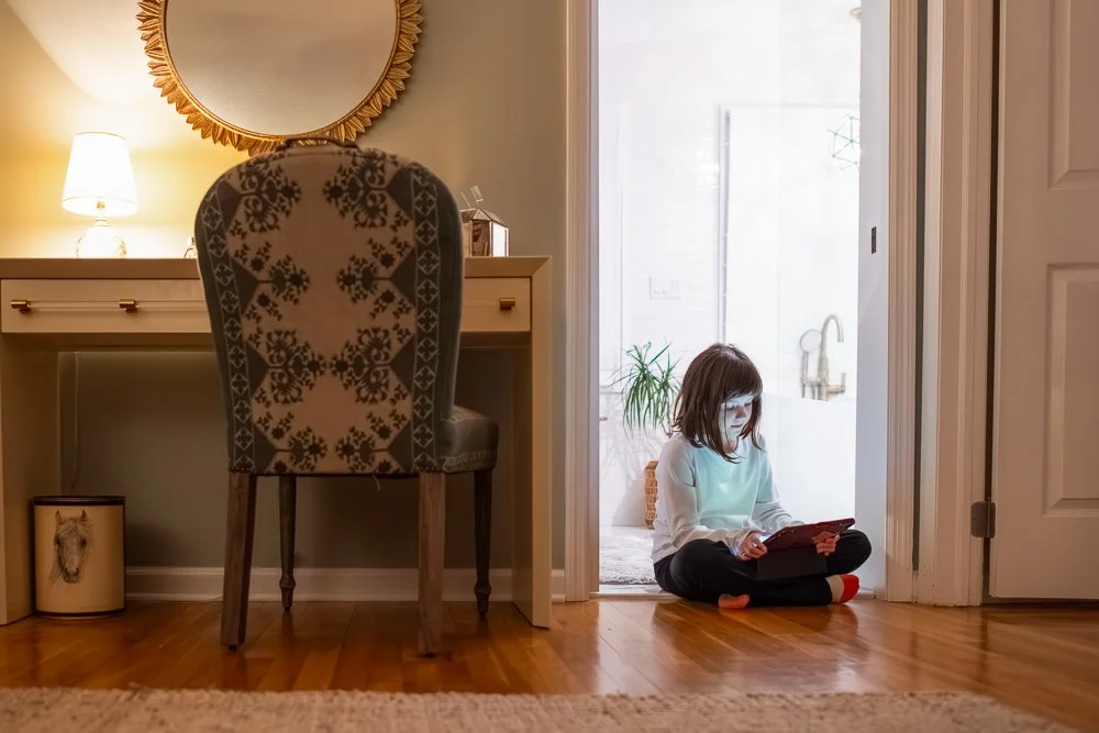 A young girl sits on the floor with her back against the doorway, looking at a device that glows. The room is dimly lit, with a lamp on a desk, a decorative mirror, and a horse picture on the wall.