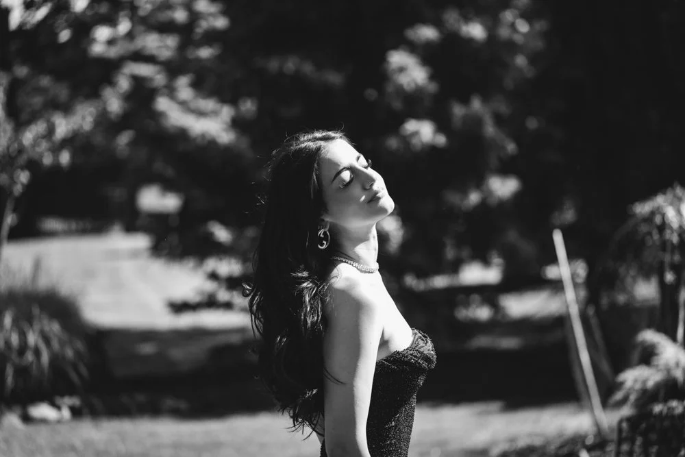 A woman with long, dark hair in a strapless dress standing outdoors with her eyes closed and face tilted upward, sunlight shining on her, surrounded by trees and grass.