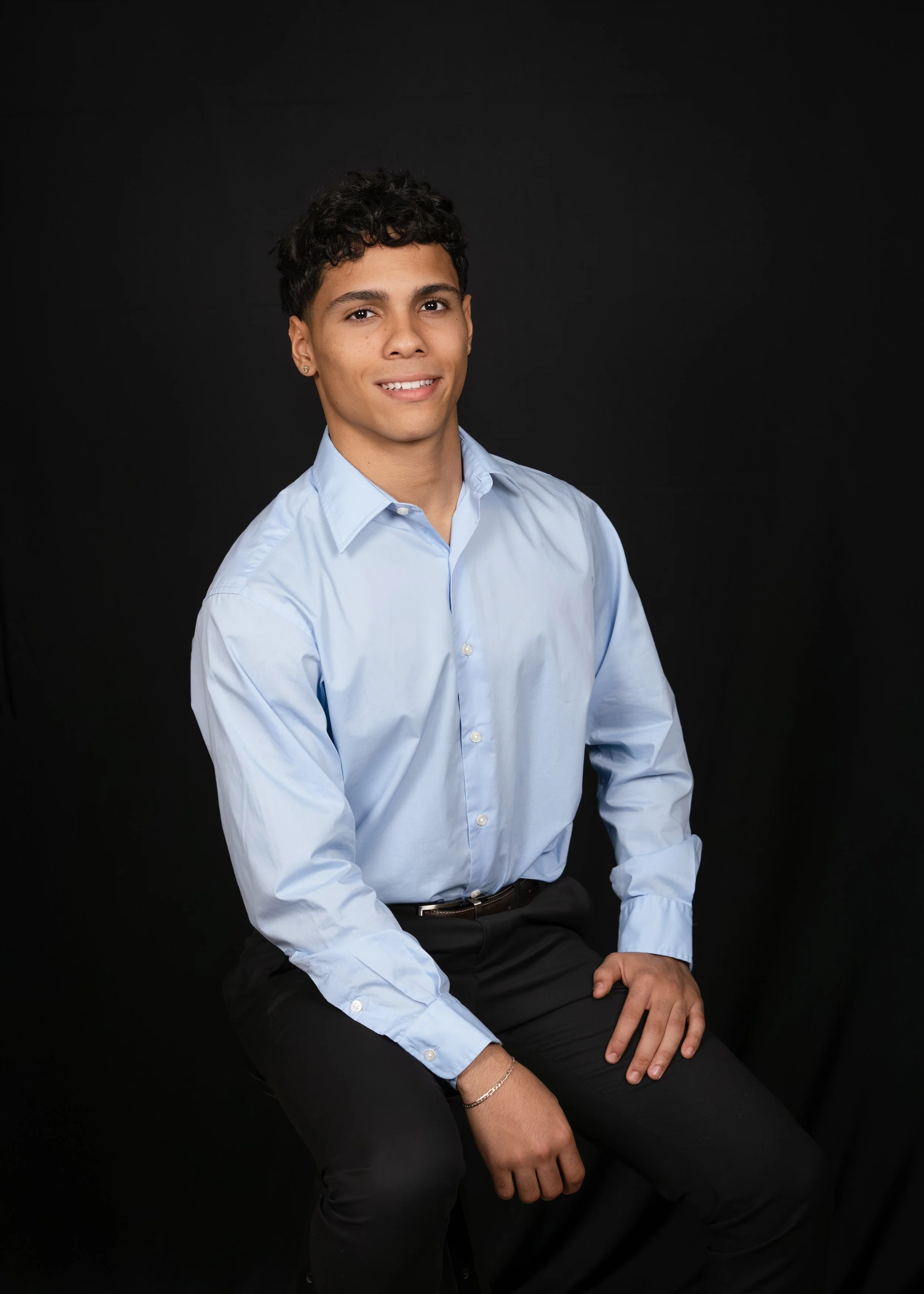 A young man with curly dark hair, wearing a light blue button-up shirt and black pants, sitting against a black background.