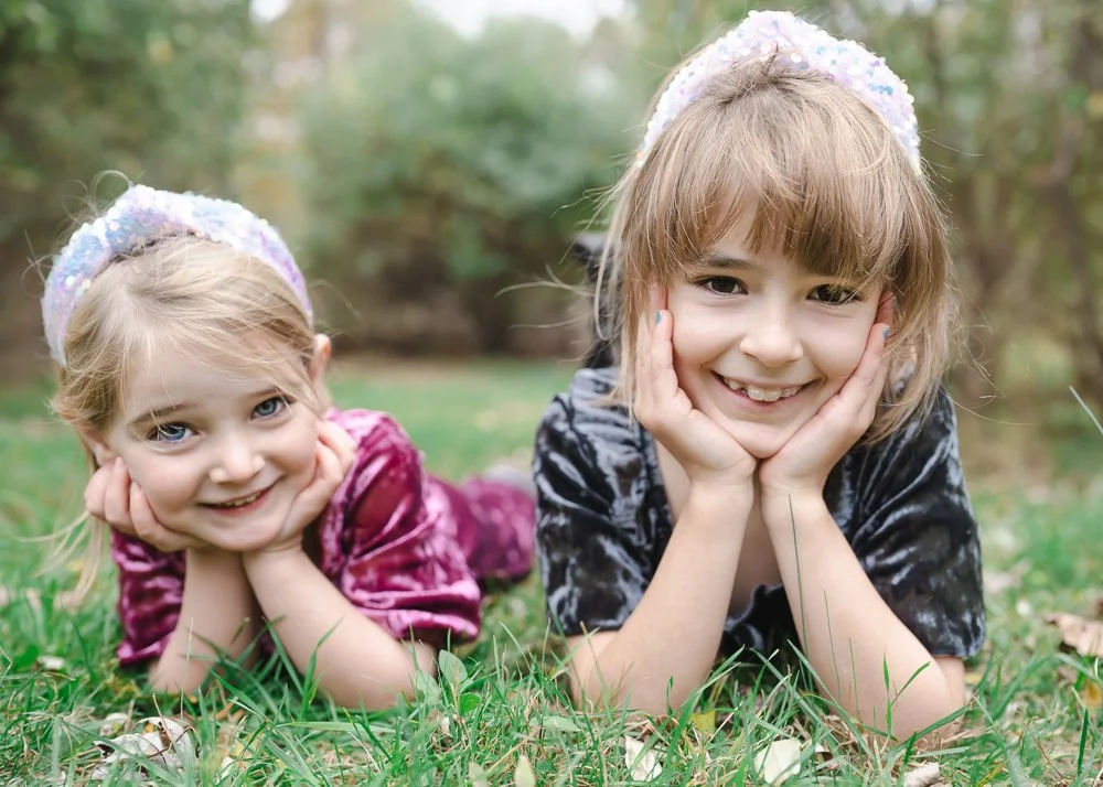 Two young girls with light brown hair and blue eyes, wearing colorful velvet dresses and sparkly headbands, lying on grass in a park or garden, smiling at the camera.