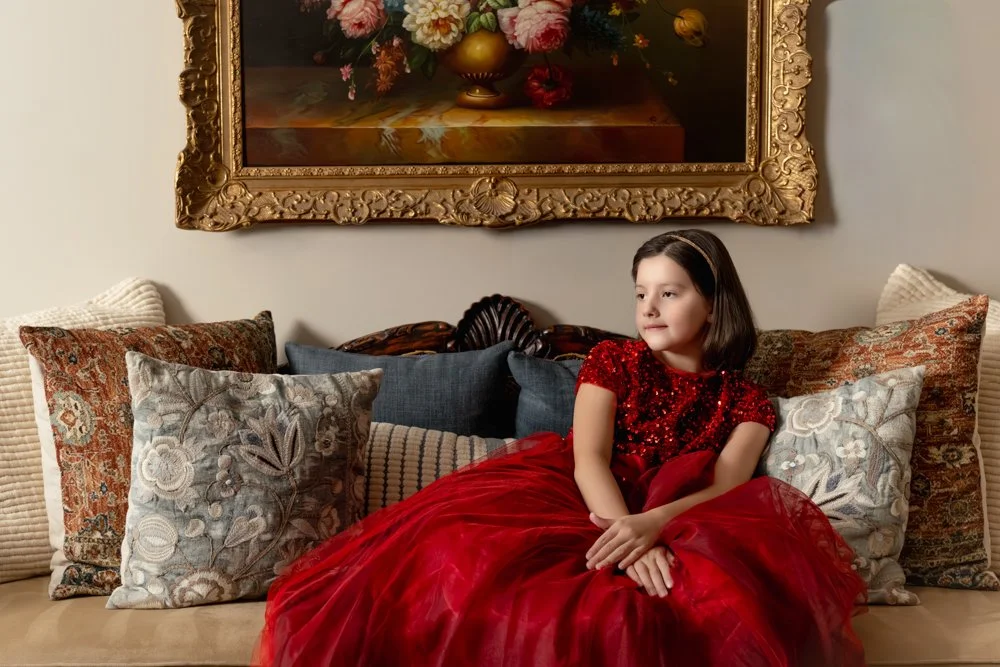 A young girl in a red dress sitting on a beige couch surrounded by decorative pillows with various patterns, in a room with a large framed floral painting on the wall behind her.