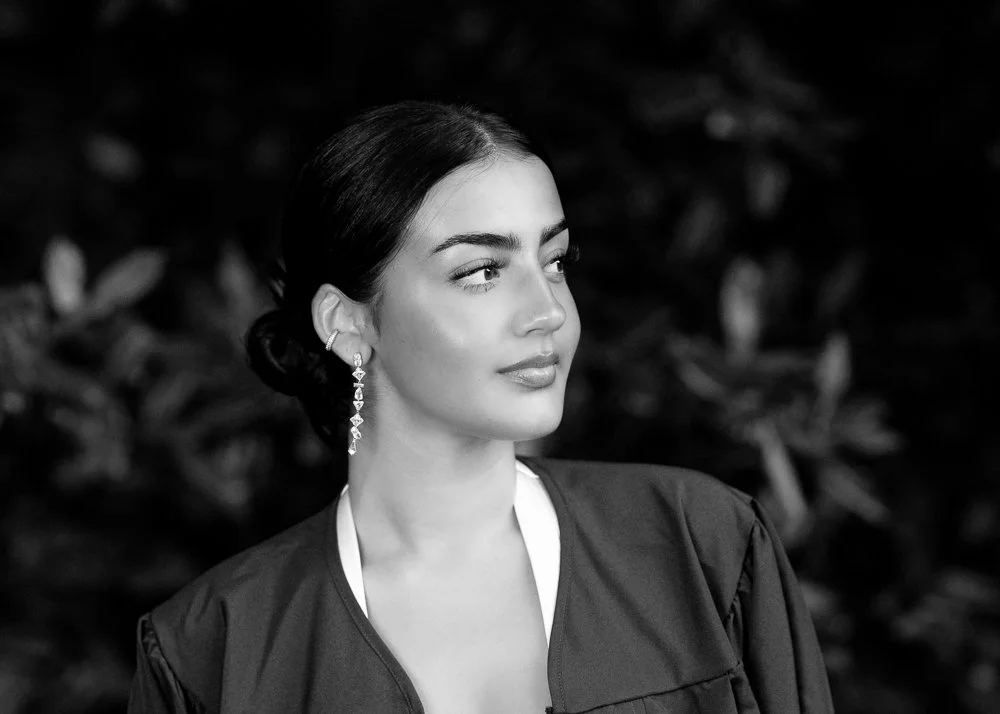 A young woman with curly dark hair and light skin, wearing a black sleeveless top, standing outdoors in a green, wooded area with blurred trees in the background.