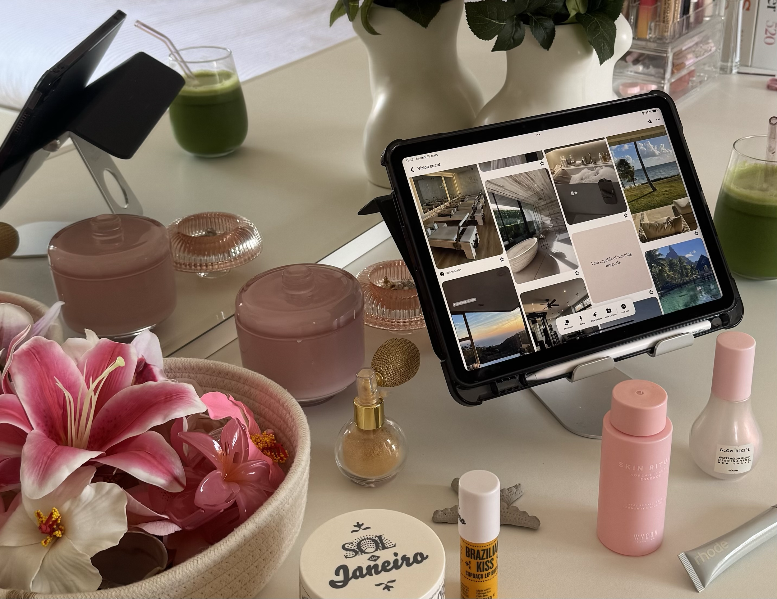 A tablet on a stand displaying a Pinterest page, surrounded by beauty and decor products including pink roses in a white vase, candles, skincare bottles, a jewelry dish, and perfume bottles on a white desk.