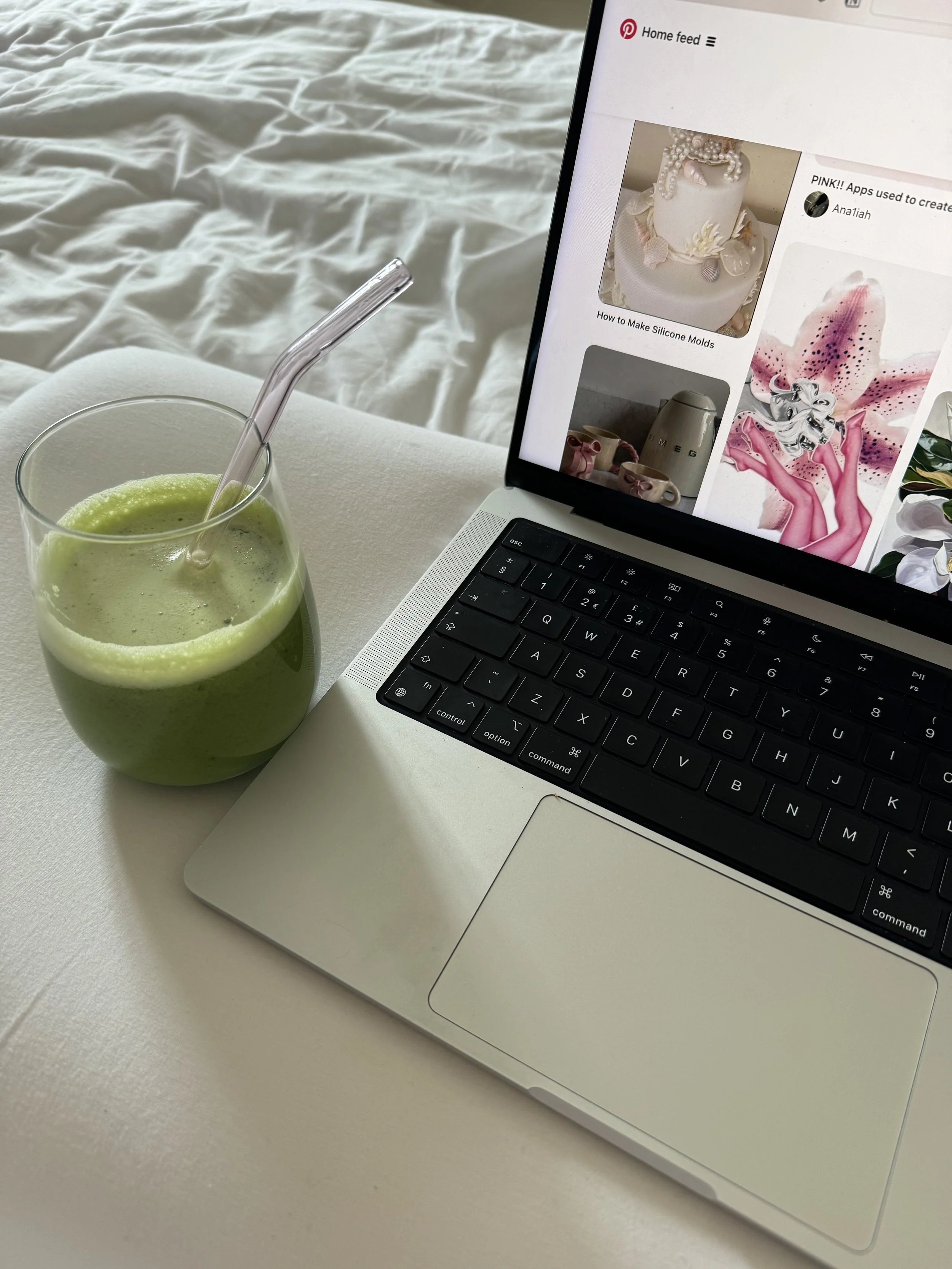 A glass of green smoothie with a straw sits on a white surface next to a silver laptop displaying Pinterest, with a background of a bed with white bedsheets.