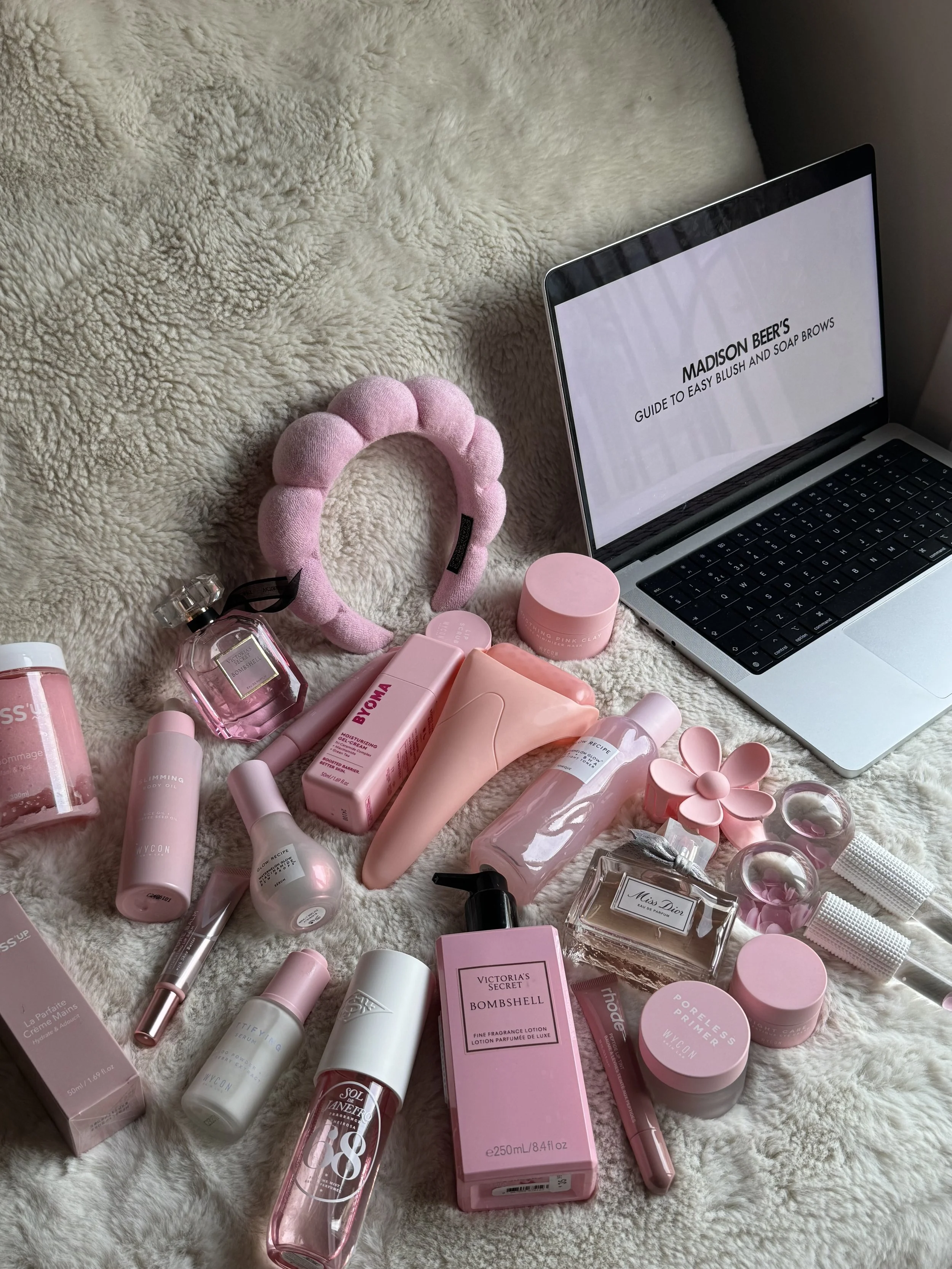 A collection of pink skincare, beauty, and self-care products spread out on a plush cream-colored blanket, with a pink fluffy headband, a pink flower-shaped beauty tool, and an open laptop displaying 'Madison Beer’s Guide to Easy Blush and Soap Brows' across the screen.