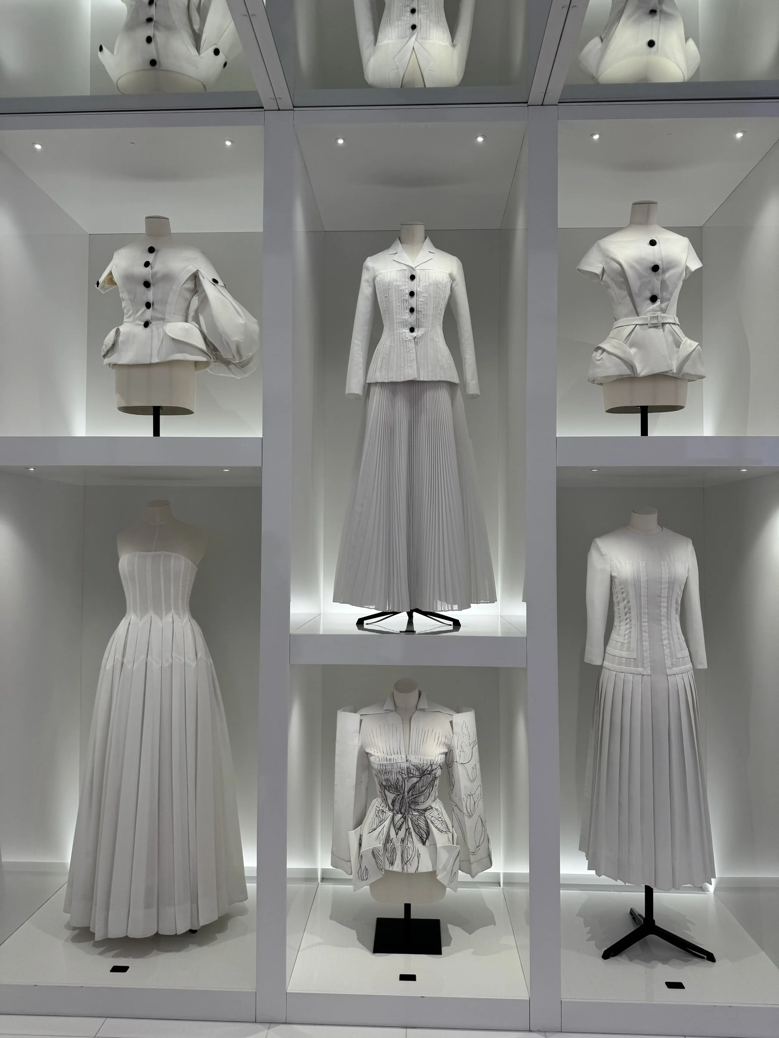 Display of white fashion dresses and jackets on mannequins in a store window with bright lighting in Paris Luxury Museum.