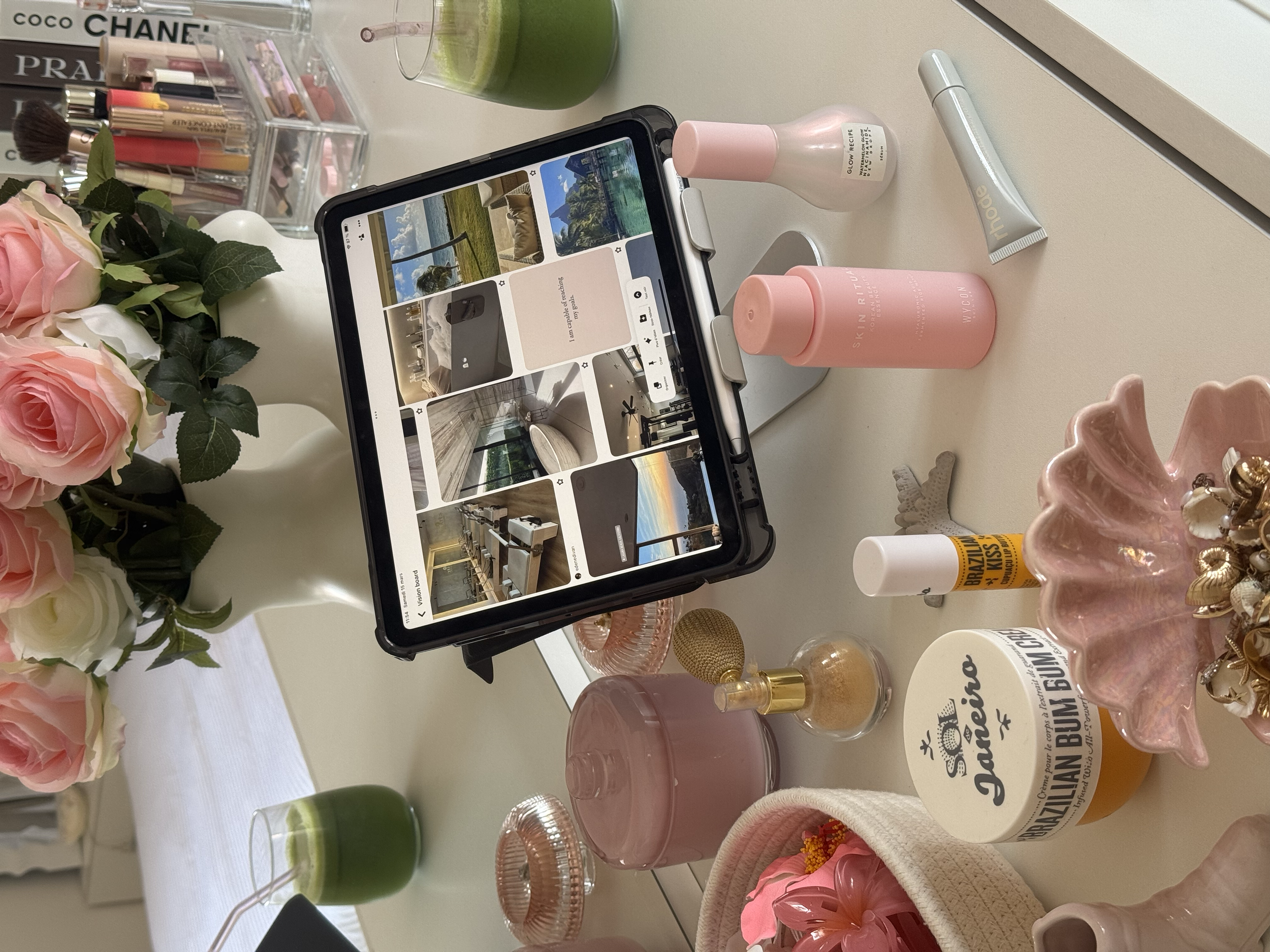 A tablet on a stand displaying a Pinterest page, surrounded by beauty and decor products including pink roses in a white vase, candles, skincare bottles, a jewelry dish, and perfume bottles on a white desk.