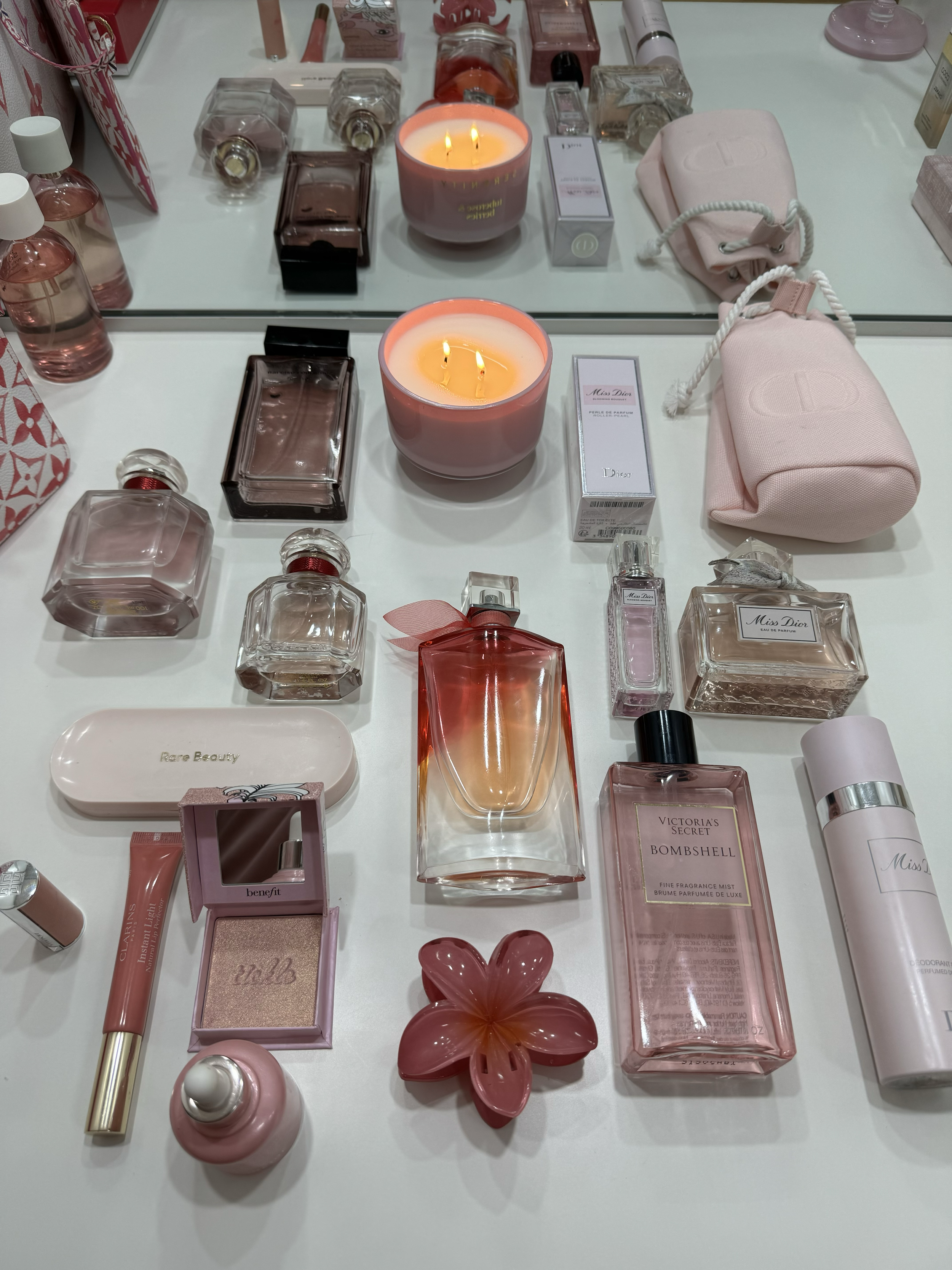 A display of various perfume bottles, skincare products, a pink candle with lit flames, a pink floral decor item, and pink pouches on a white surface with a mirror reflecting the items. Pinterest style.