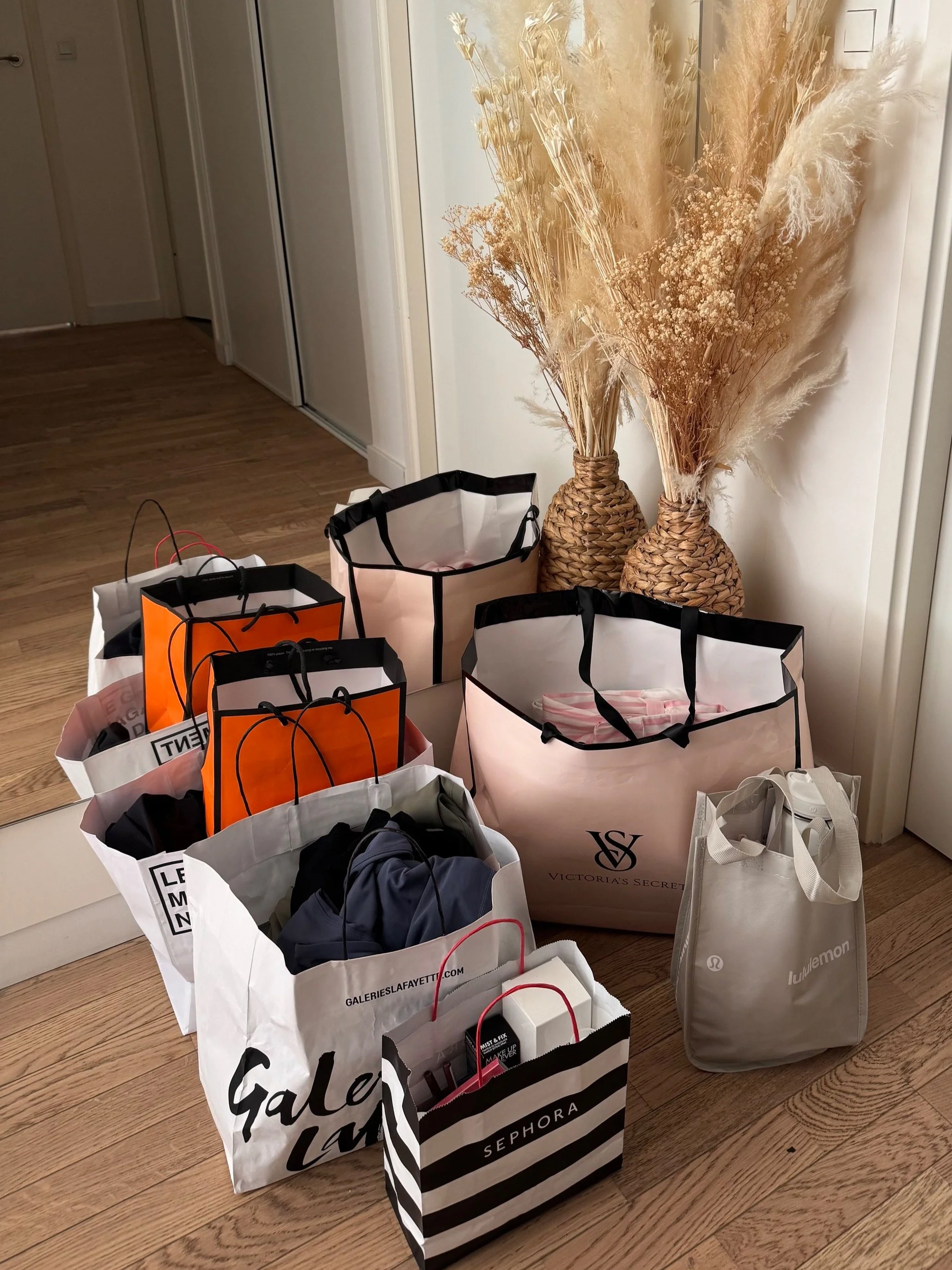 Multiple shopping bags on a wooden floor near two woven vases with dried pampas grass and flowers.