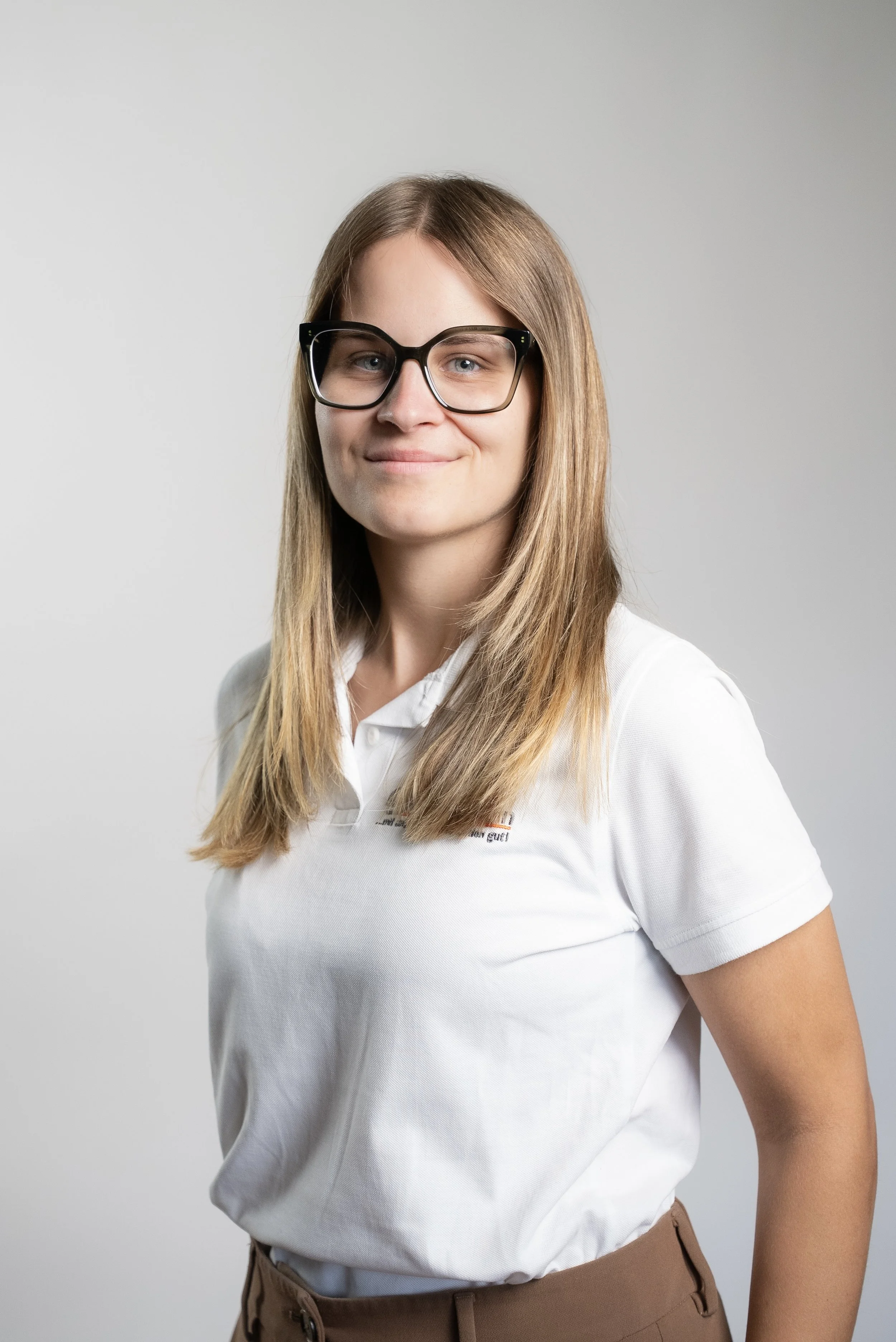 Junge Frau mit Brille und braunen Haaren, trägt ein weißes Polo-Shirt, lächelt leicht, steht vor hellem Hintergrund.