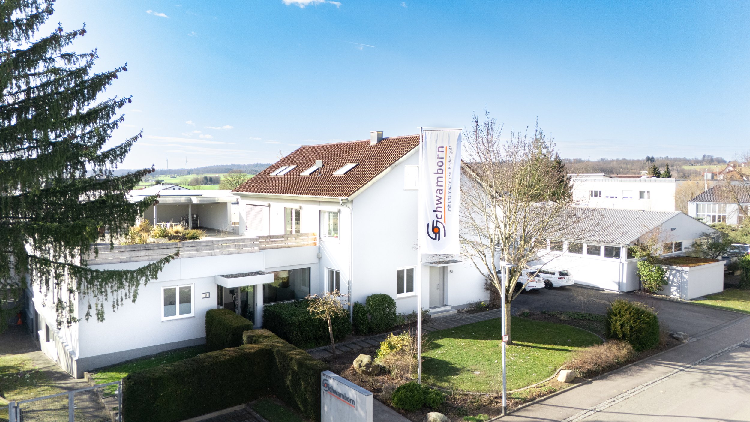 Ein modernes weißes Gebäude mit mehreren Stockwerken und einem roten Ziegeldach. Es gibt Bäume, einen gepflegten Rasen und geparkte Autos. Eine Fahne mit dem Logo und dem Namen 'Schwamborn' weht im Wind.
