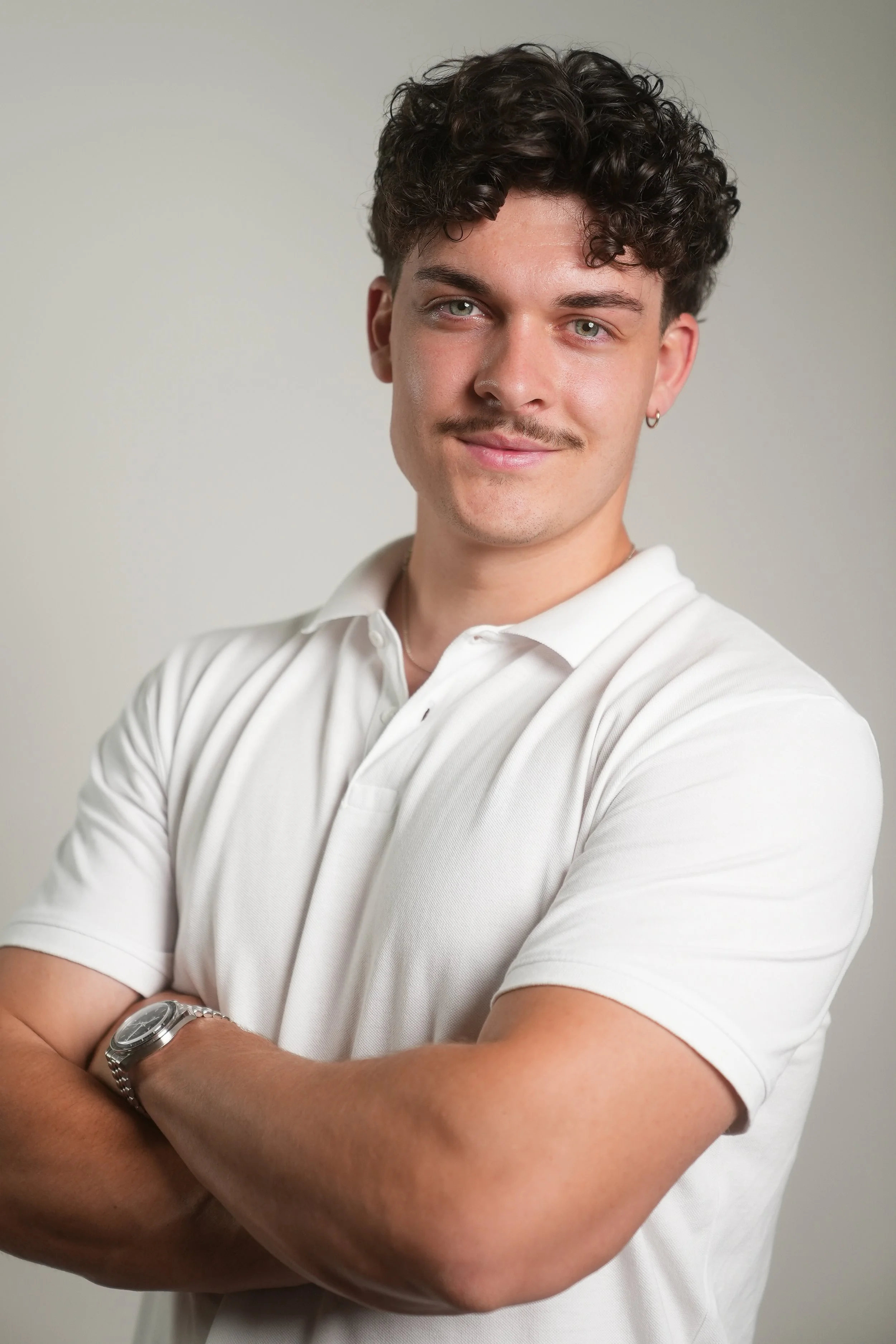 Junger Mann mit hellen Haaren und leichter Tönungsmustache trägt weißen Polo-Shirt, trägt Armbanduhr, steht vor neutralem Hintergrund, lächelt leicht, mit verschränkten Armen.