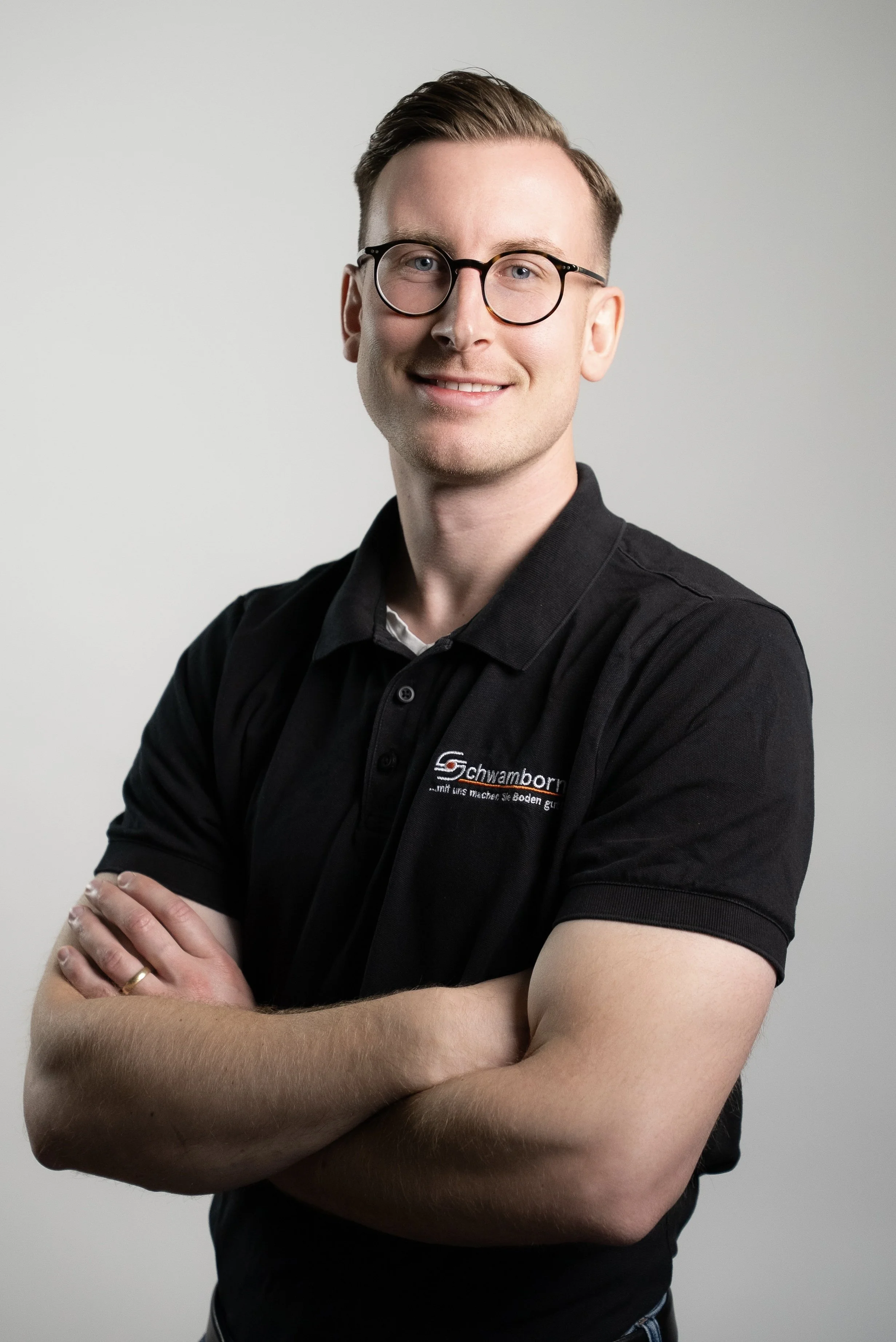 Ein junger Mann mit Brille, kurz freundlich lächelnd, trägt ein schwarzes Polo-Shirt mit einem Logo, steht vor hellem Hintergrund, Arme verschränkt.