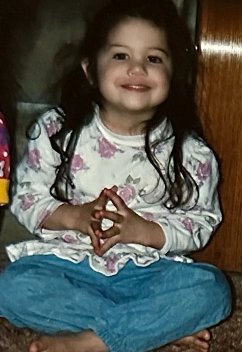 Young Brittany Mondido with long dark hair, sitting cross-legged on the floor, smiling and clasping her hands together in mudra, wearing a floral long-sleeve shirt and blue pants.