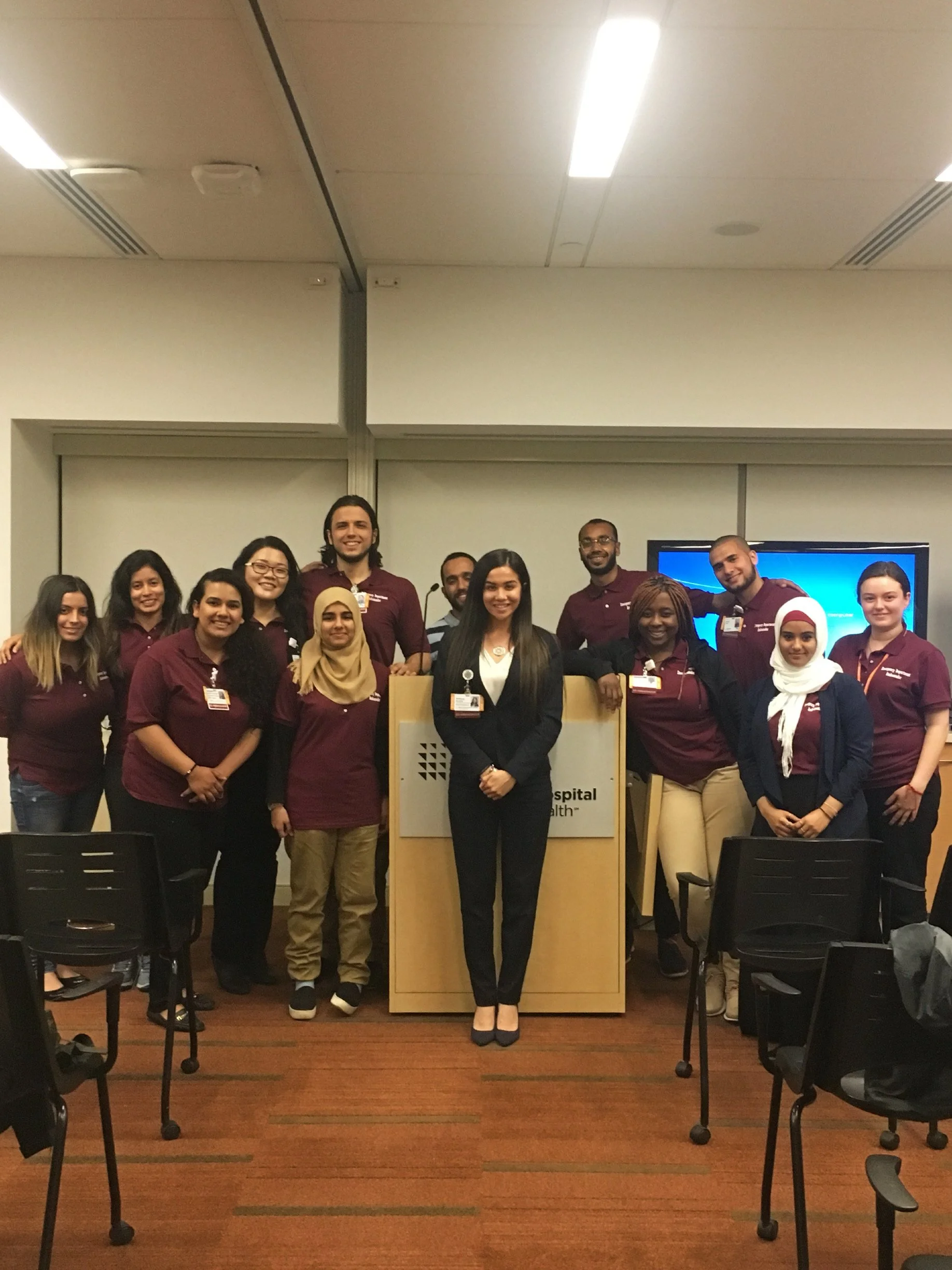 Group of diverse healthcare professionals and students standing together in a conference room wearing maroon shirts, and Brittany Mondido, thought leader, in a black suit standing at the center in front of a podium leading the Northwell Health team.