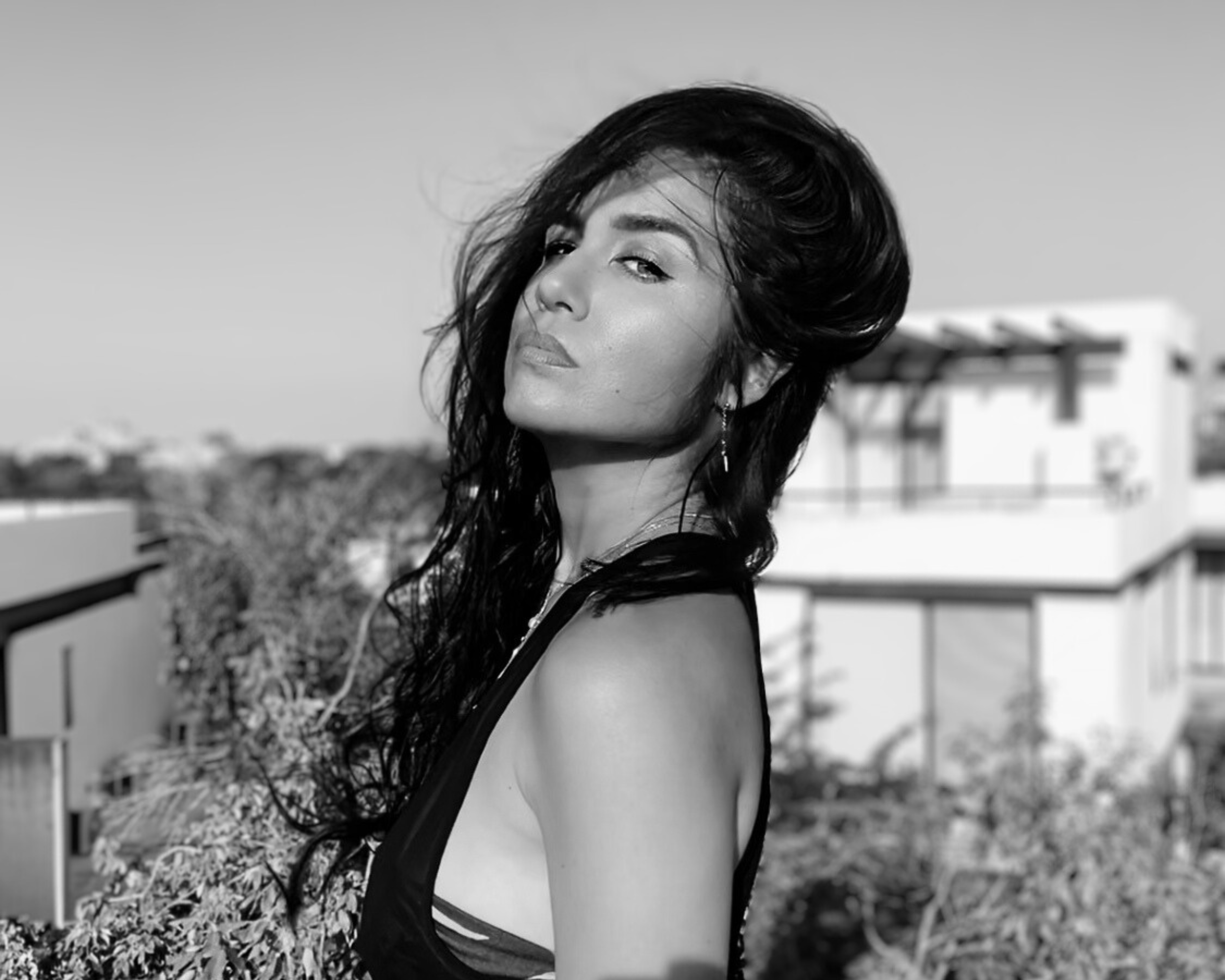Black and white photo of a woman with long, wavy hair standing outdoors on a rooftop, with buildings and trees in the background.