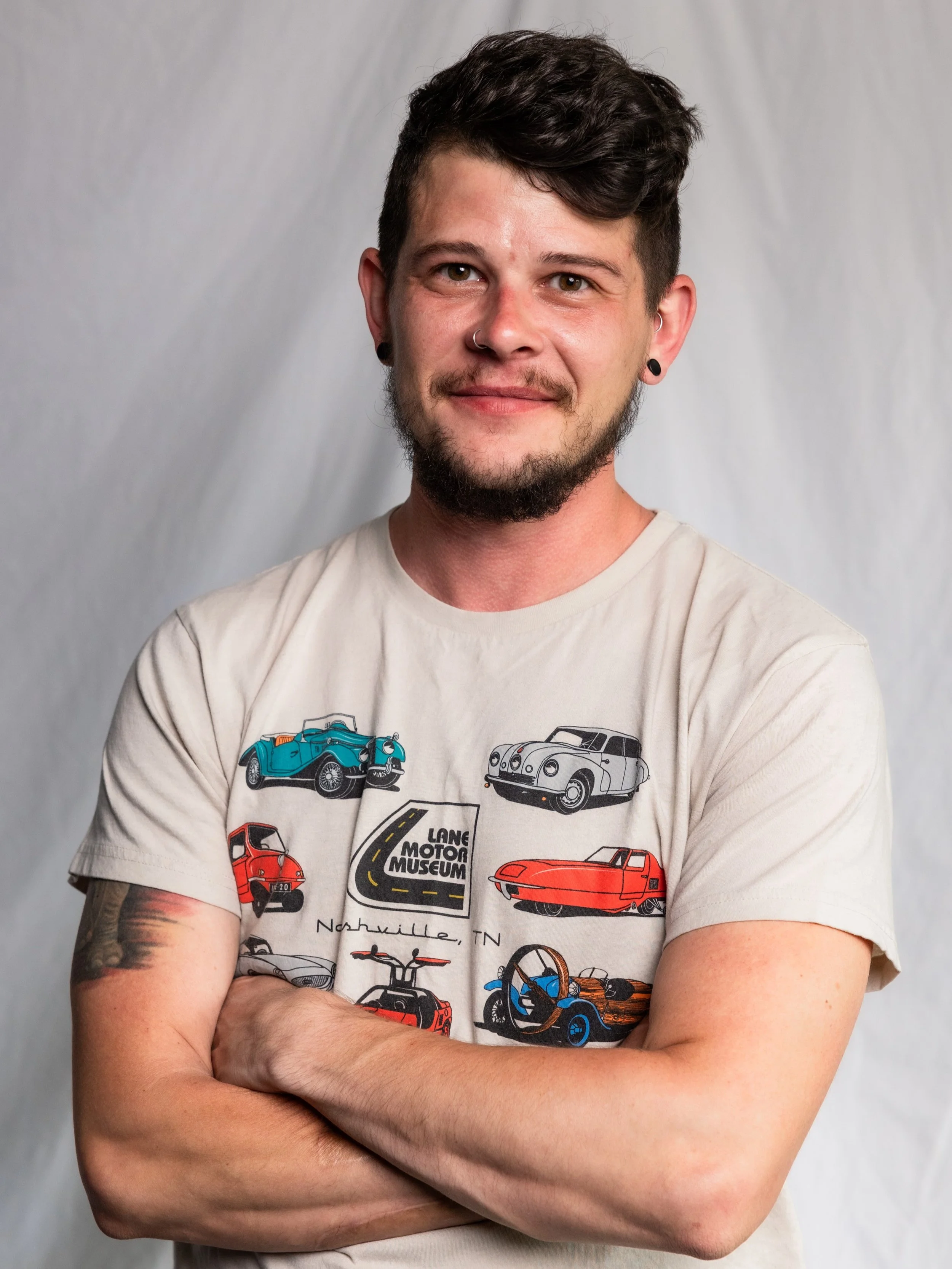 A young man with dark hair, beard, and tattoos, smiling with arms crossed, wearing a beige t-shirt with vintage cars and the text 'Lane Motor Museum Nashville TN'.